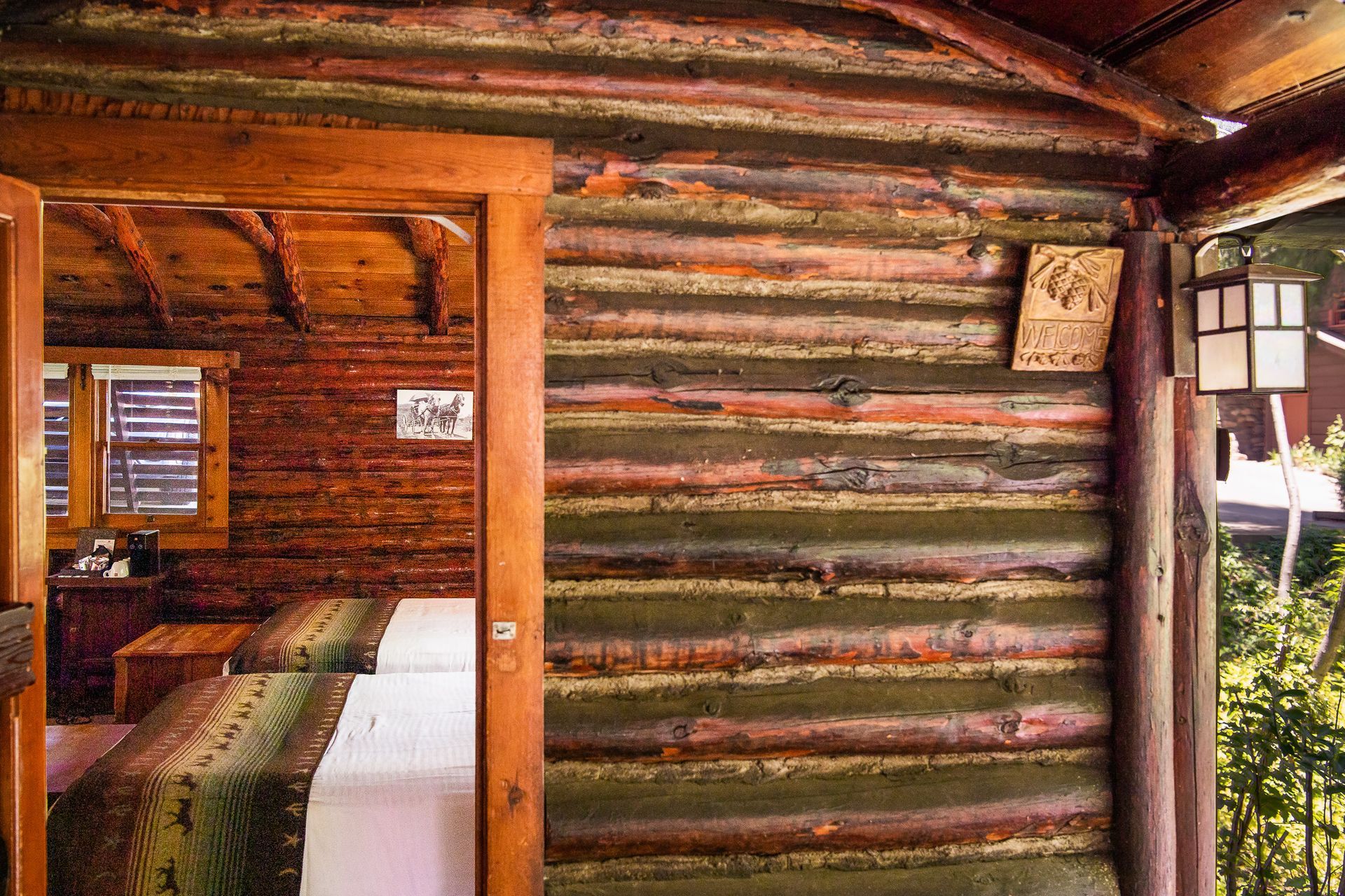 Wooden cabin interior; bedroom visible through doorway with bed and window. Brown, green, and red hues.