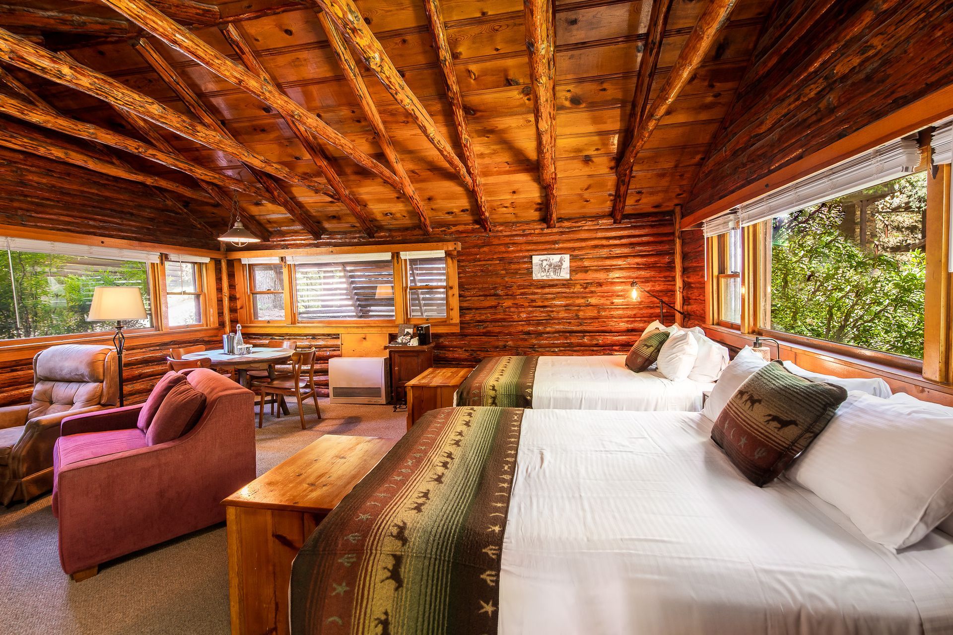 Rustic cabin bedroom with two beds, armchair, and small table; wooden walls and ceiling.