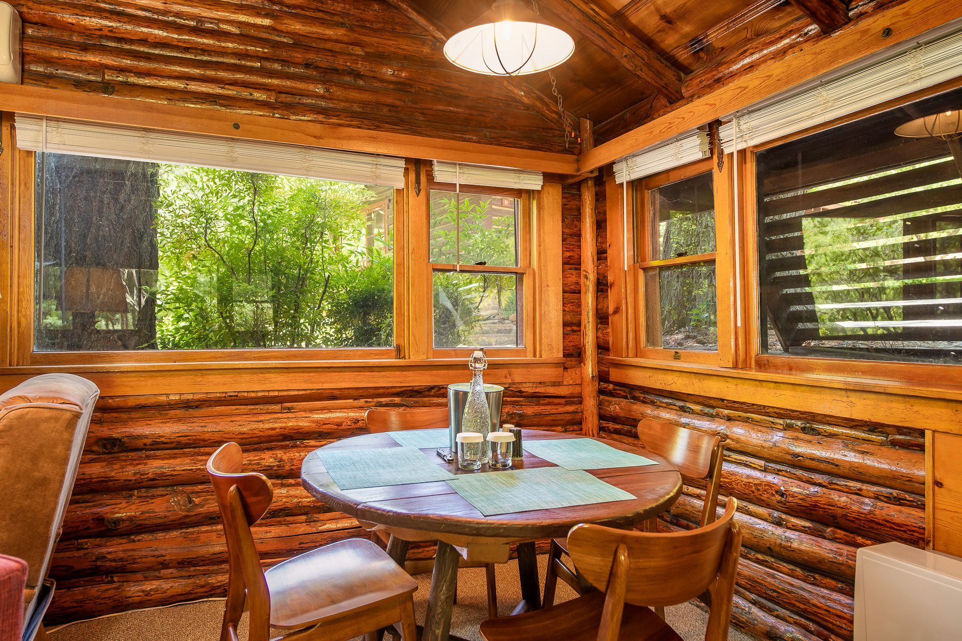 Cozy cabin interior: wooden walls, round table with four chairs, windows overlooking lush greenery.