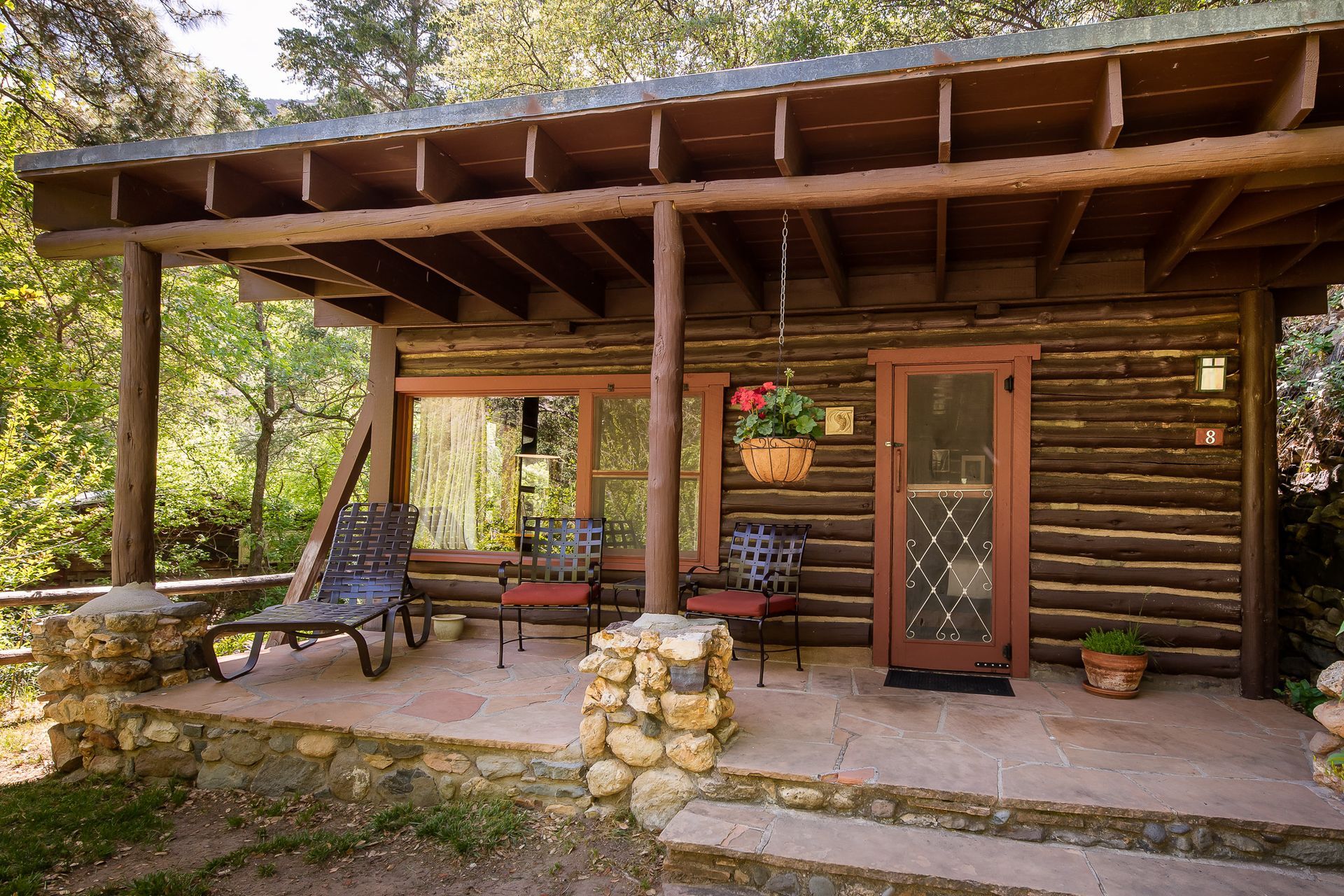 Log cabin with porch, chairs, and hanging flower basket. Rustic exterior with stone and wood elements.