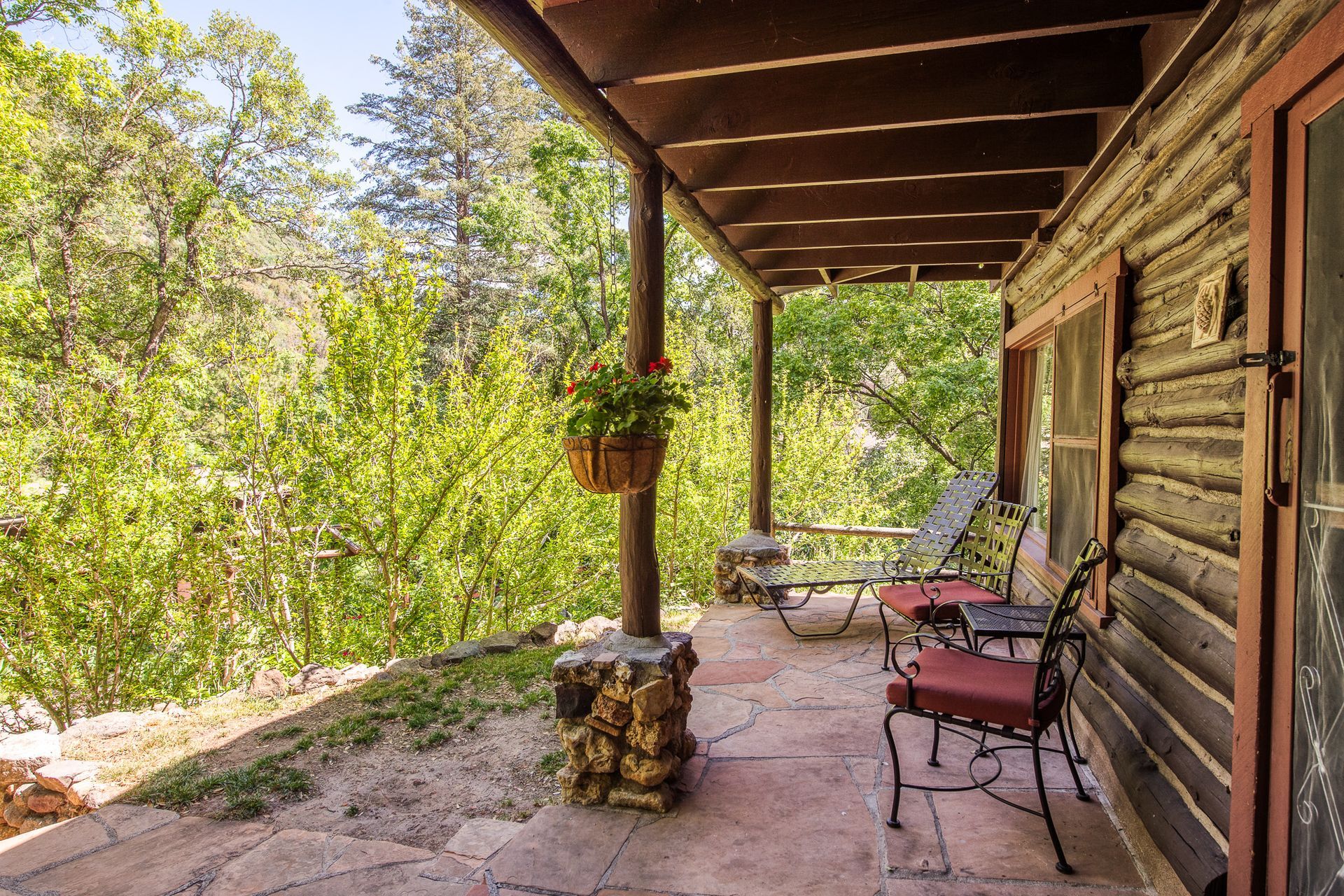 Covered porch of a rustic cabin with chairs, plants, and a view of trees.