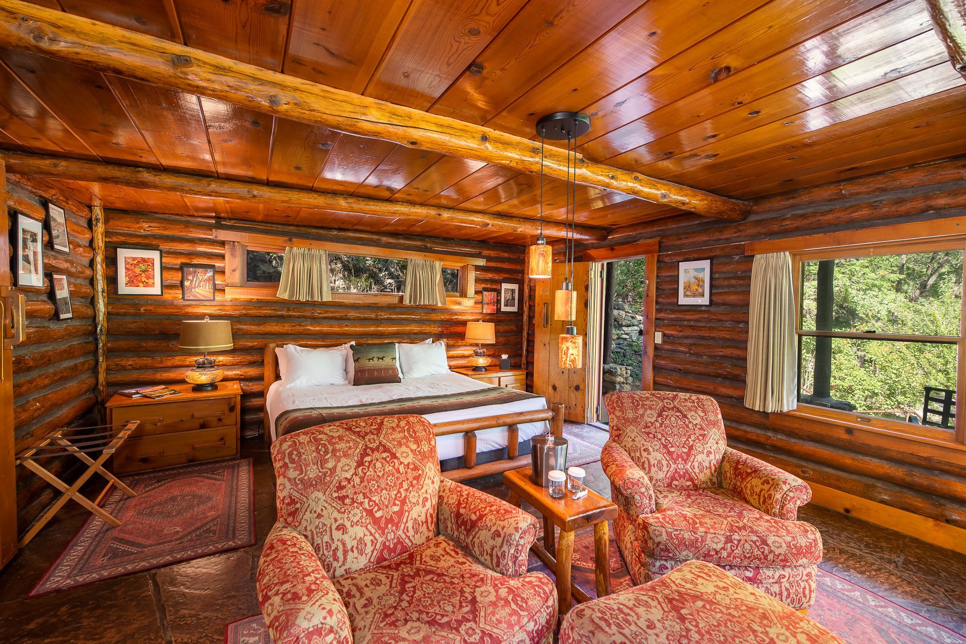 Log cabin bedroom with wood paneling, a bed, patterned armchairs, and a window with a forest view.