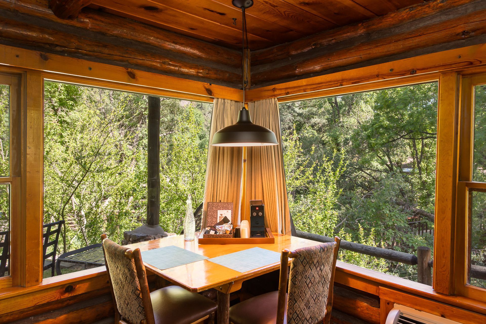 Cozy dining nook with a table and two chairs, windows overlooking lush greenery, and a pendant light.