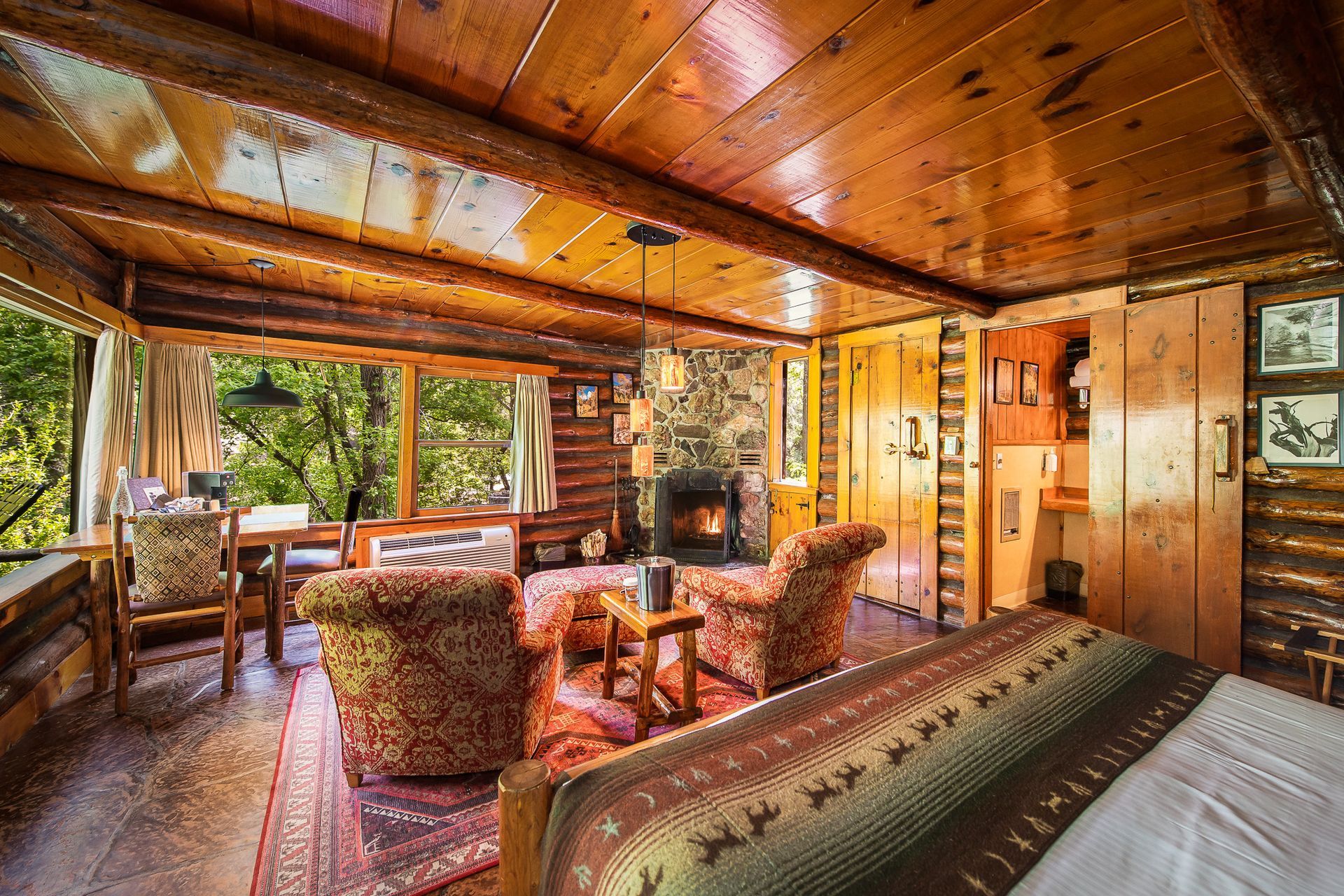 Rustic cabin interior: wooden walls/ceiling, fireplace, two armchairs, bed, and window view of trees.