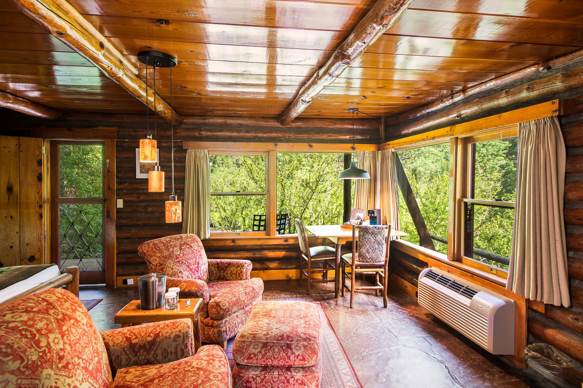 Cozy cabin interior with log walls, two armchairs, a small table, and large windows overlooking lush greenery.