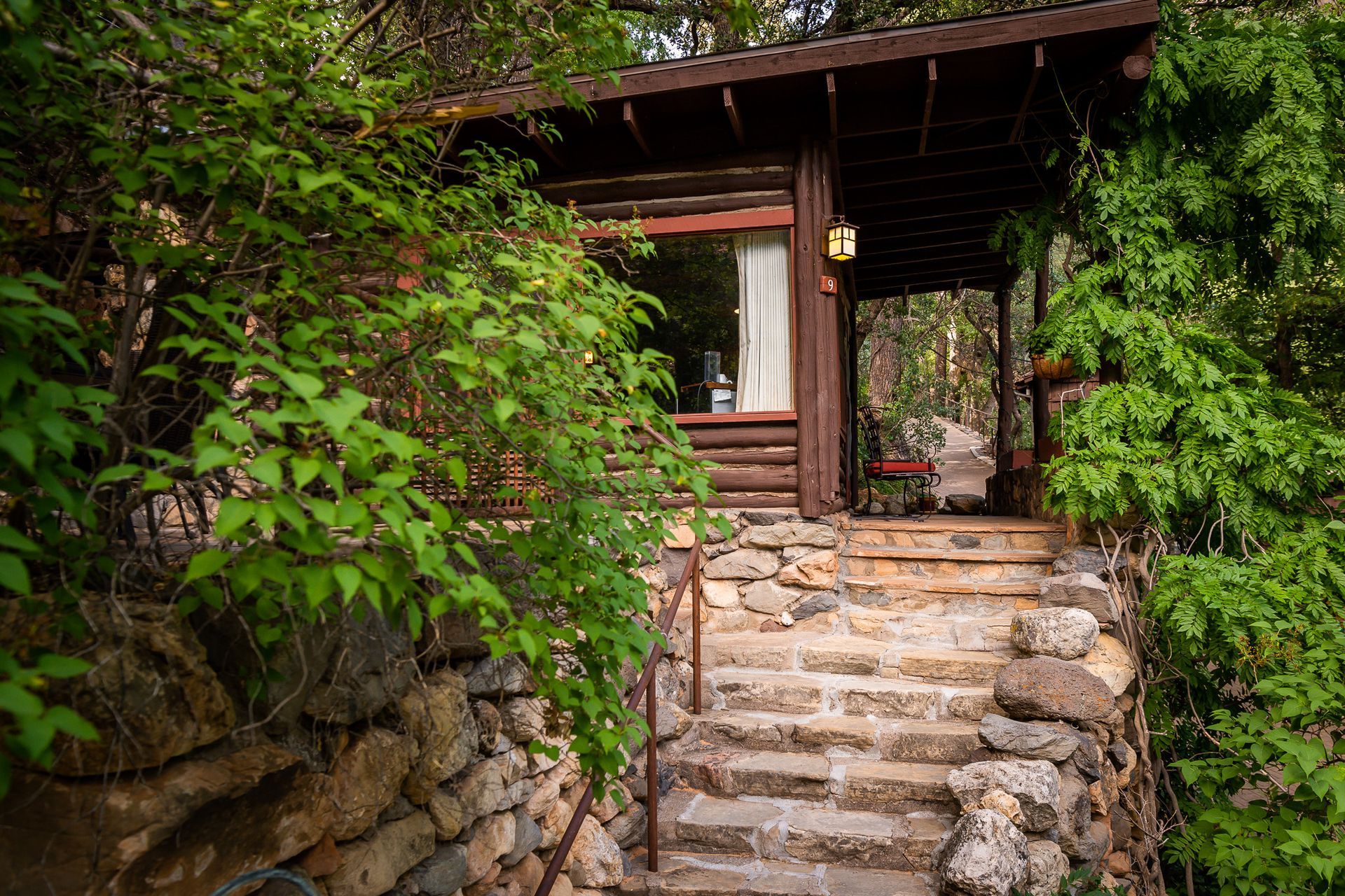 Stone steps lead to a wooden cabin entrance covered in greenery; a small window is visible.