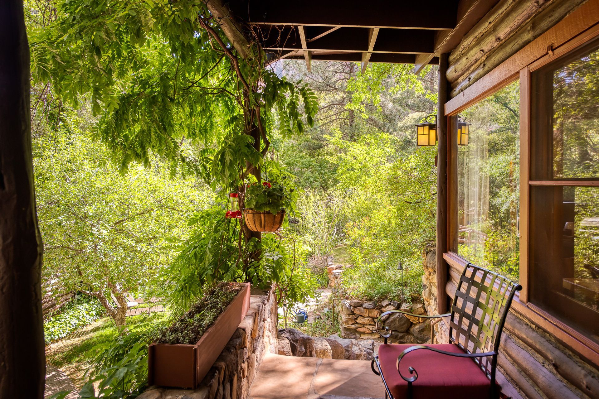 Porch with red bench and hanging flowers, surrounded by lush green trees.