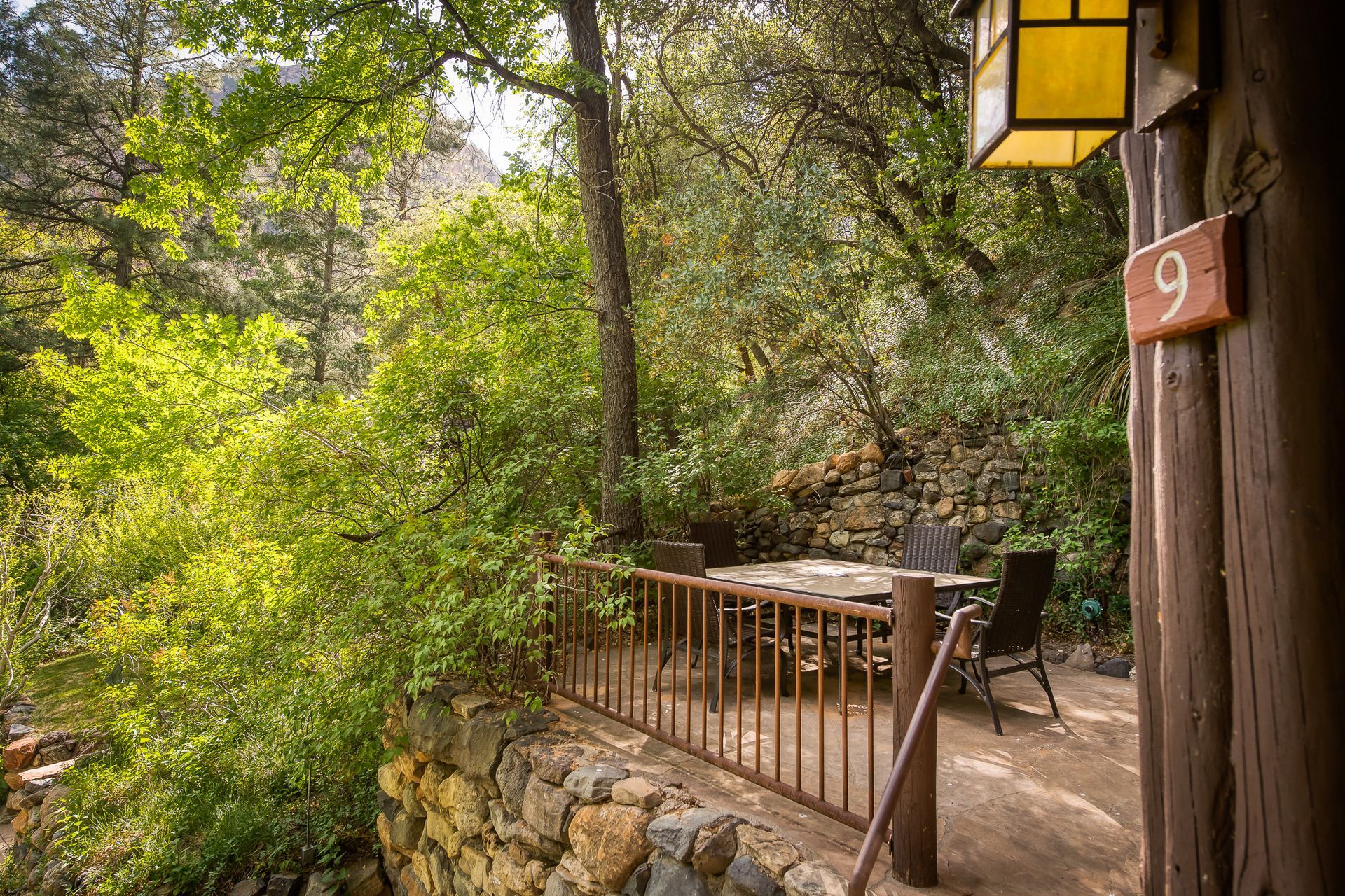 Patio of cabin 9 overlooking a lush, green forest. Brown wood, stone wall, and a table with chairs.