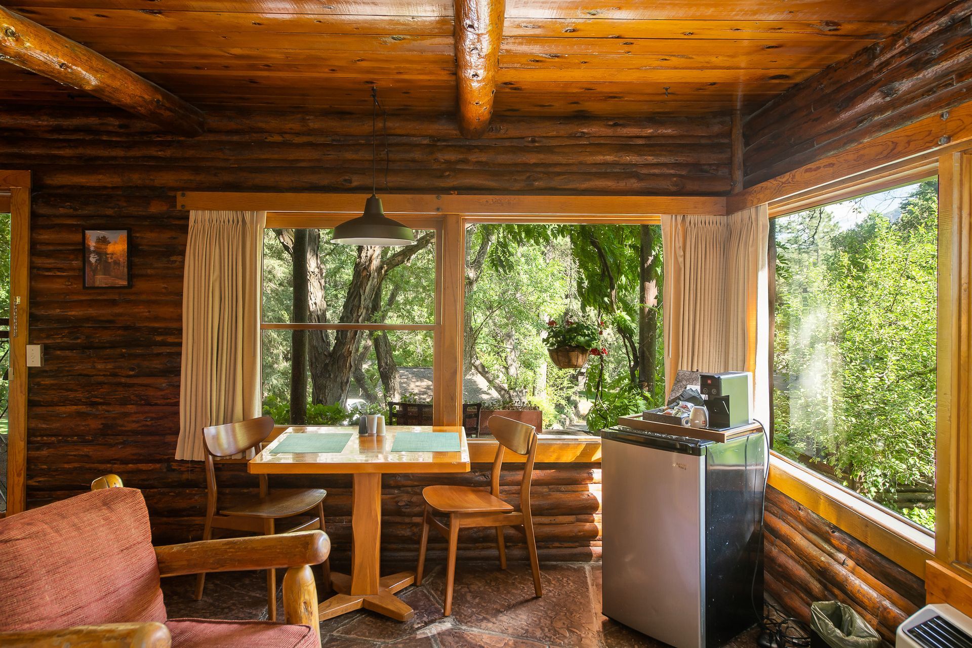 Cozy cabin interior with wooden walls, table set by a window overlooking lush trees.
