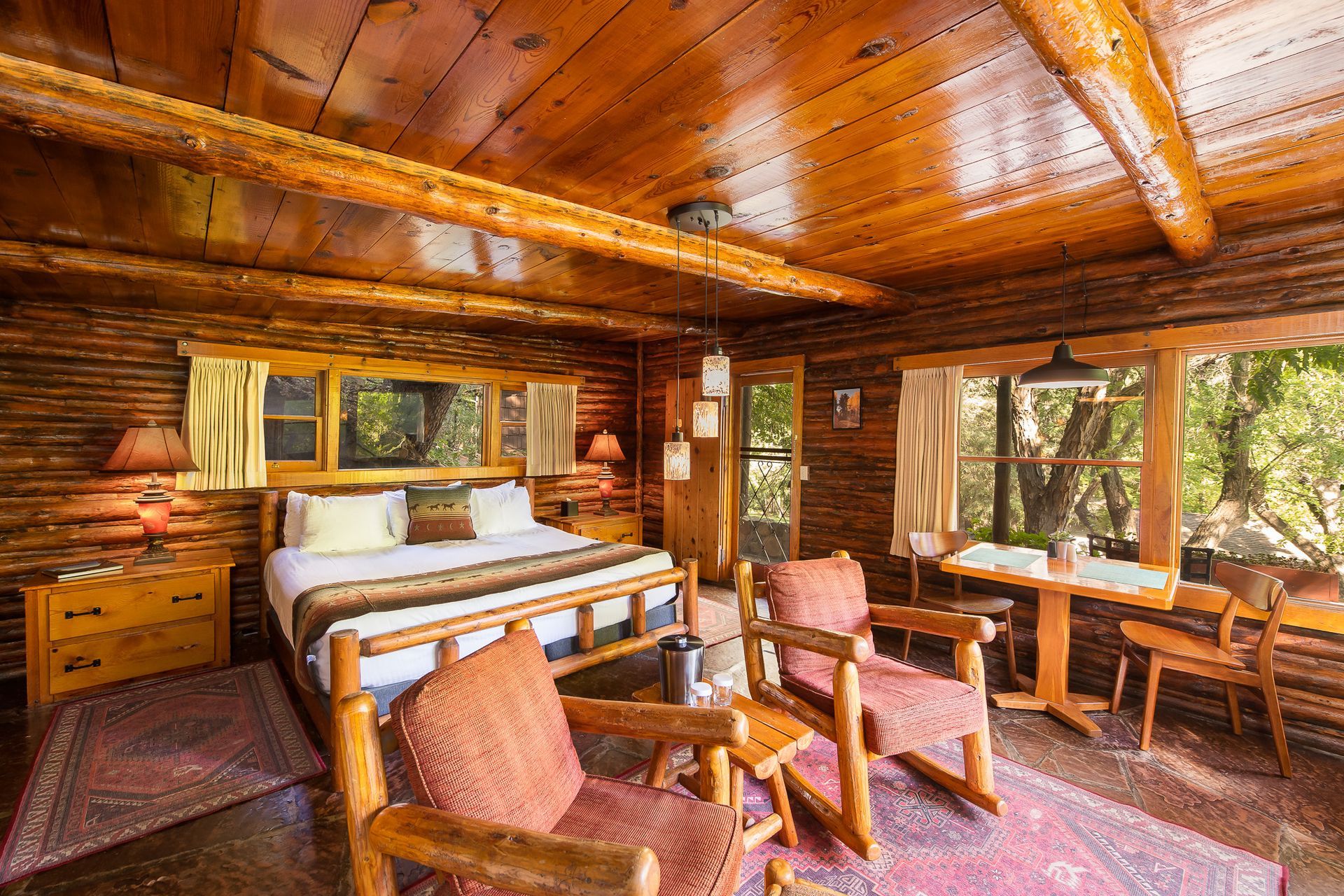 Cozy log cabin bedroom with bed, chairs, and a small table by a window overlooking nature.
