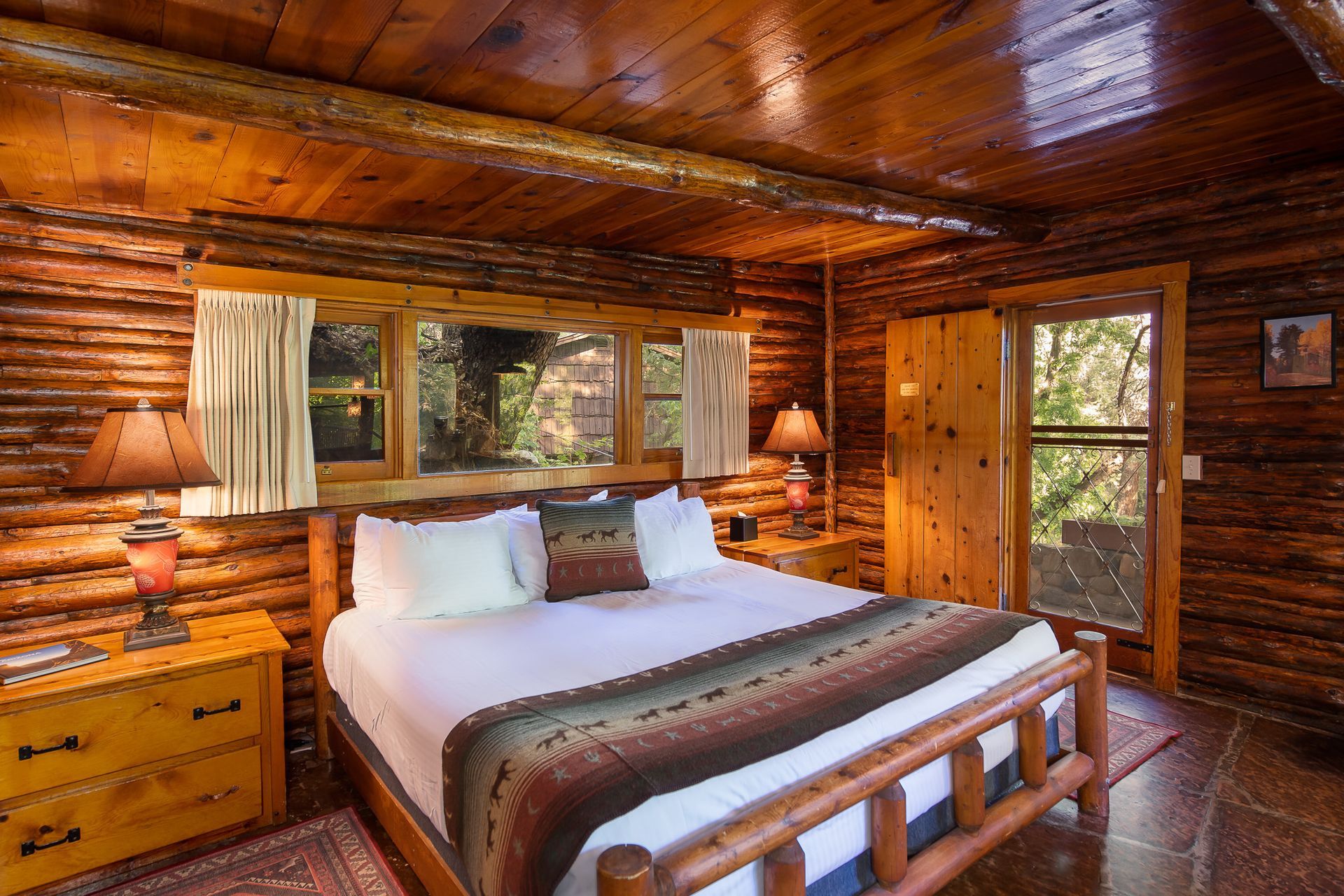 Cozy log cabin bedroom with a bed, dresser, lamps, and a window looking outside.