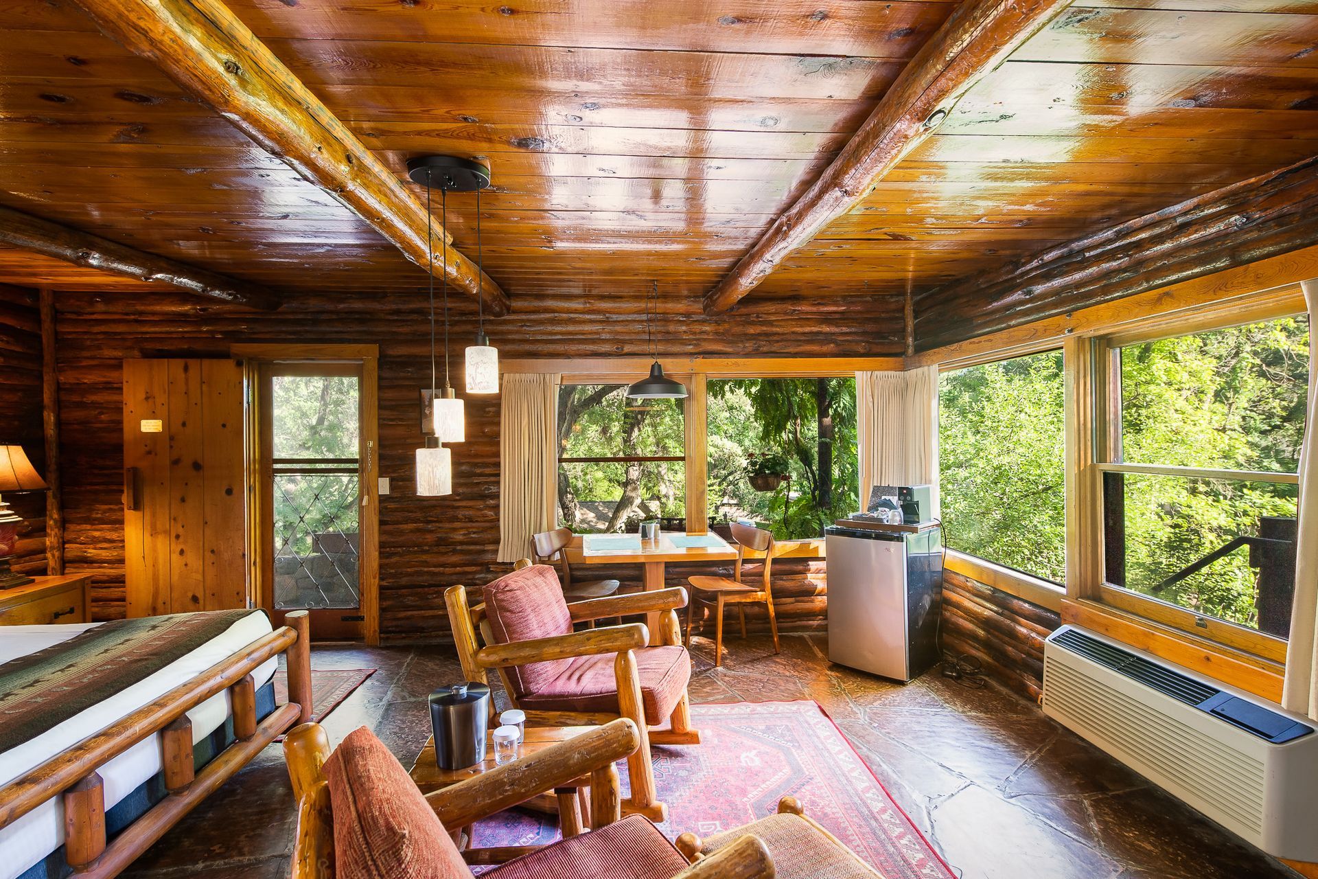 Cozy cabin interior with wood walls and ceiling, large windows looking out at trees, and rustic furniture.