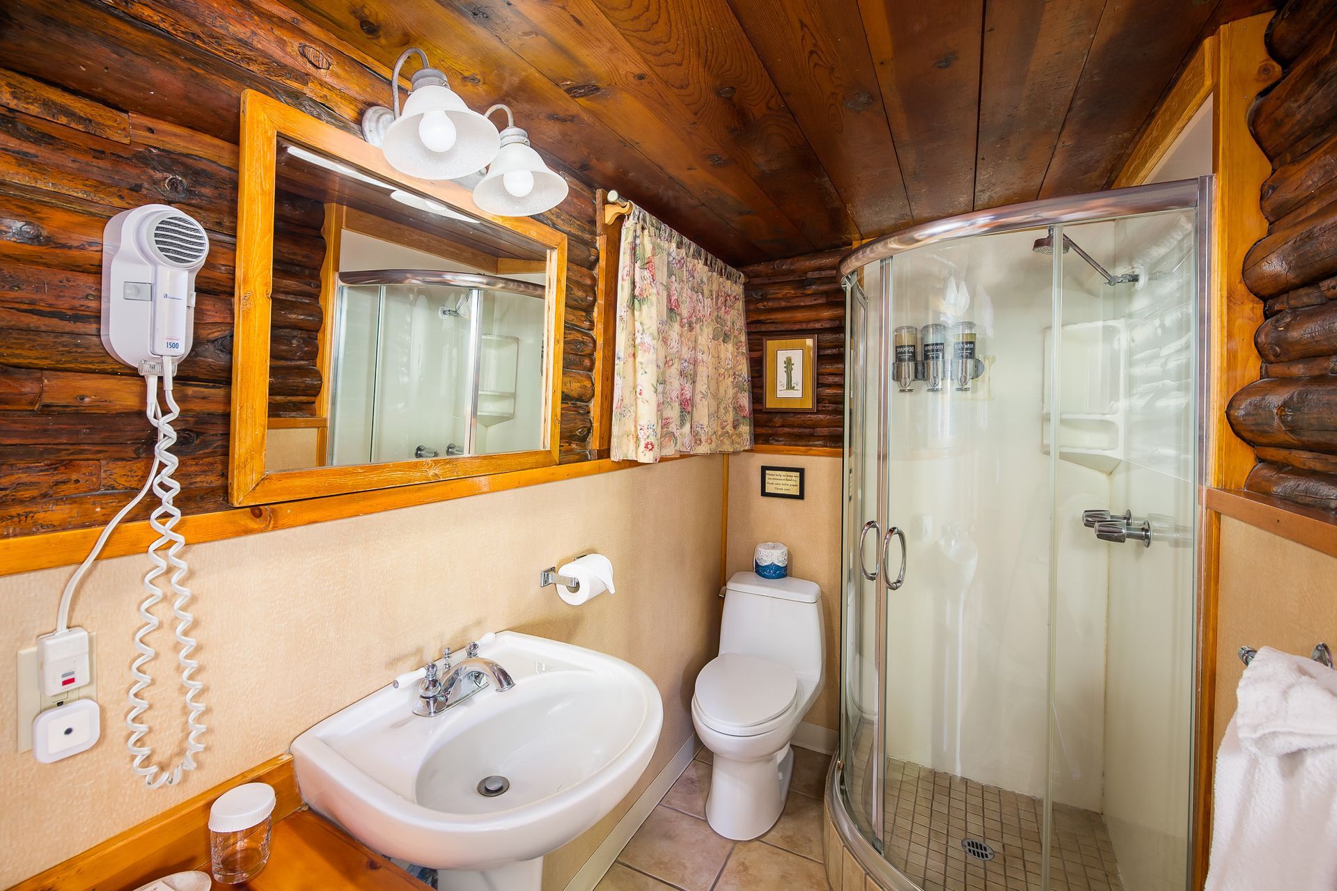Bathroom with log walls, sink, toilet, shower, and mirror. Hair dryer mounted on the wall.