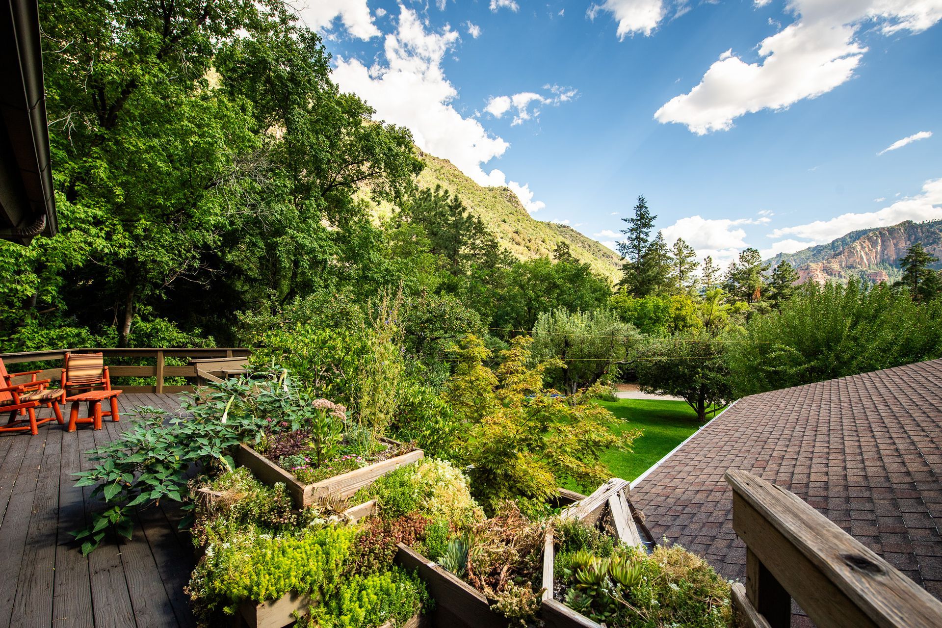 Wooden deck with gardens, overlooking lush green trees, mountains, and blue sky.