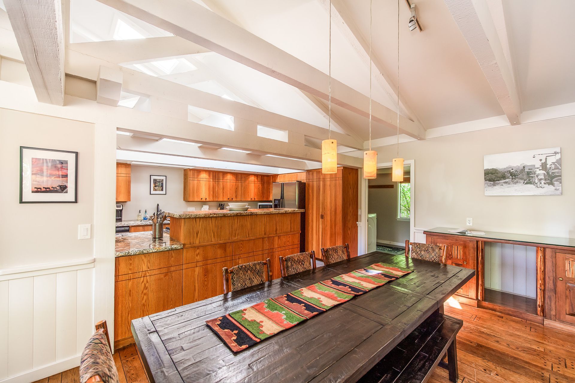 Dining room with long black table, wooden cabinets, open to a kitchen, light-colored walls, and wood floors.