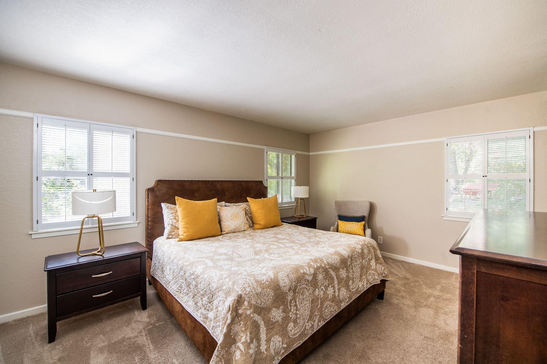 Bedroom with a bed, two windows, nightstands, and a chair. Beige walls, brown furniture, and yellow accents.