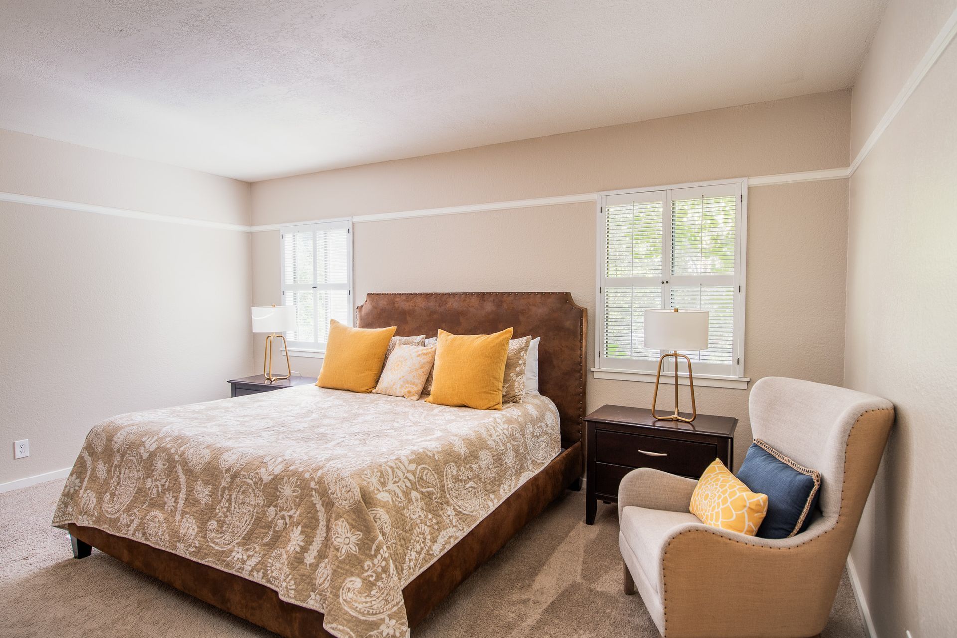 Bedroom with bed, armchair, and windows. Beige walls, patterned bedspread, and yellow pillows.