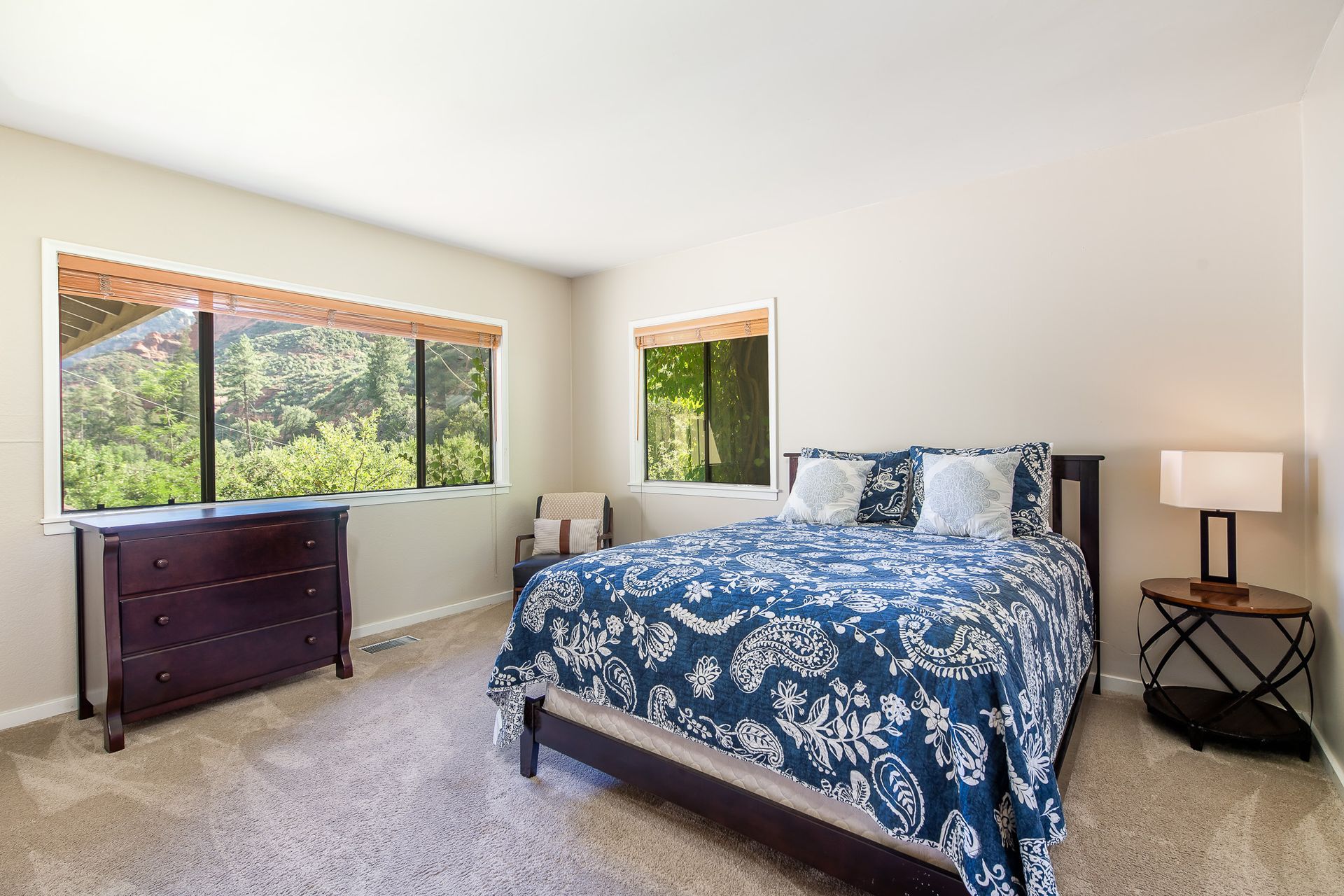 Bedroom with bed, dresser, chair, and two windows overlooking trees. Blue patterned bedspread.