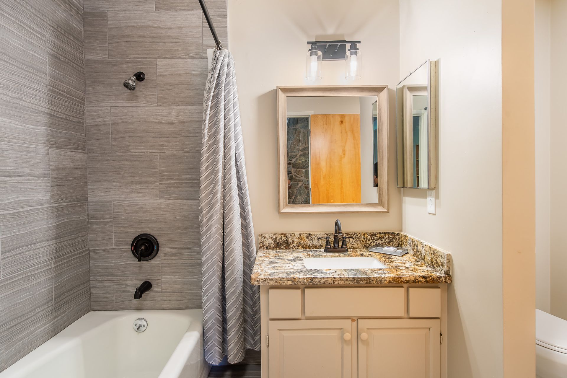 Bathroom with gray tiled shower, vanity with granite countertop, and a closed cabinet.