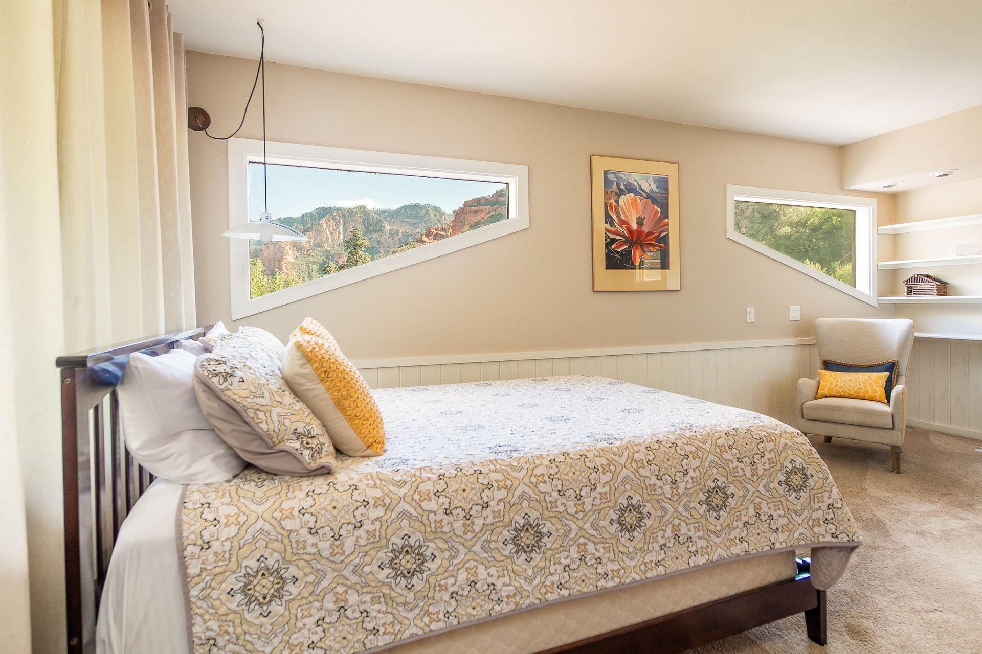 Bedroom with bed, unique windows overlooking mountains, artwork, chair, desk.