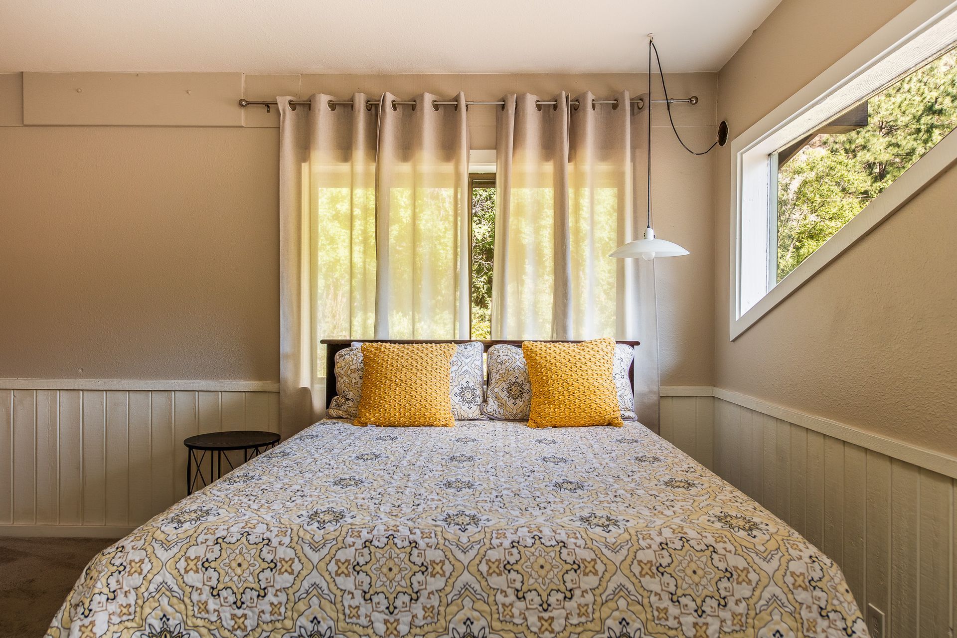 Bedroom with bed, yellow pillows, patterned quilt, window with curtains, and a pendant light.