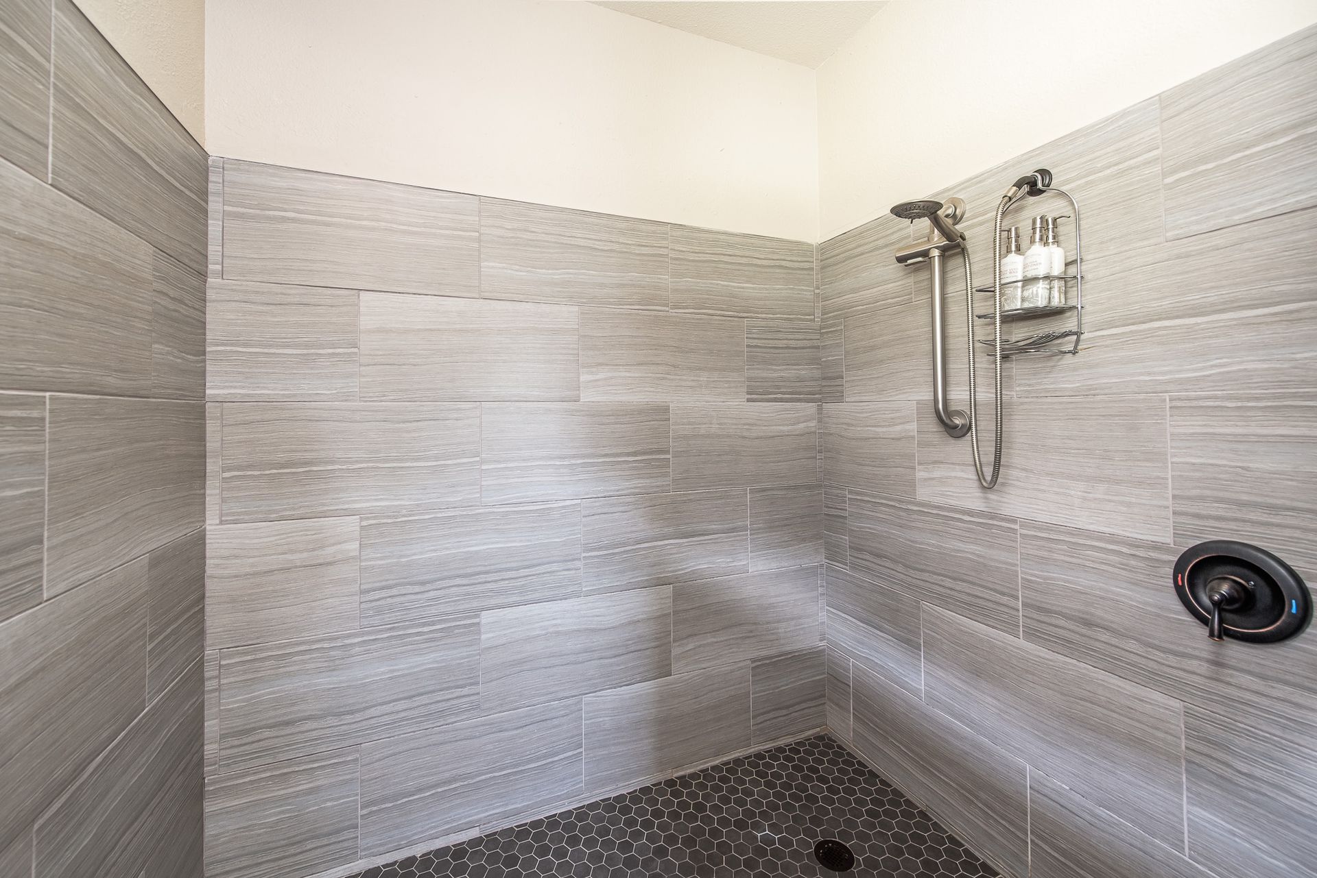 Gray tiled shower with pebble floor, shower head, and soap dispenser.