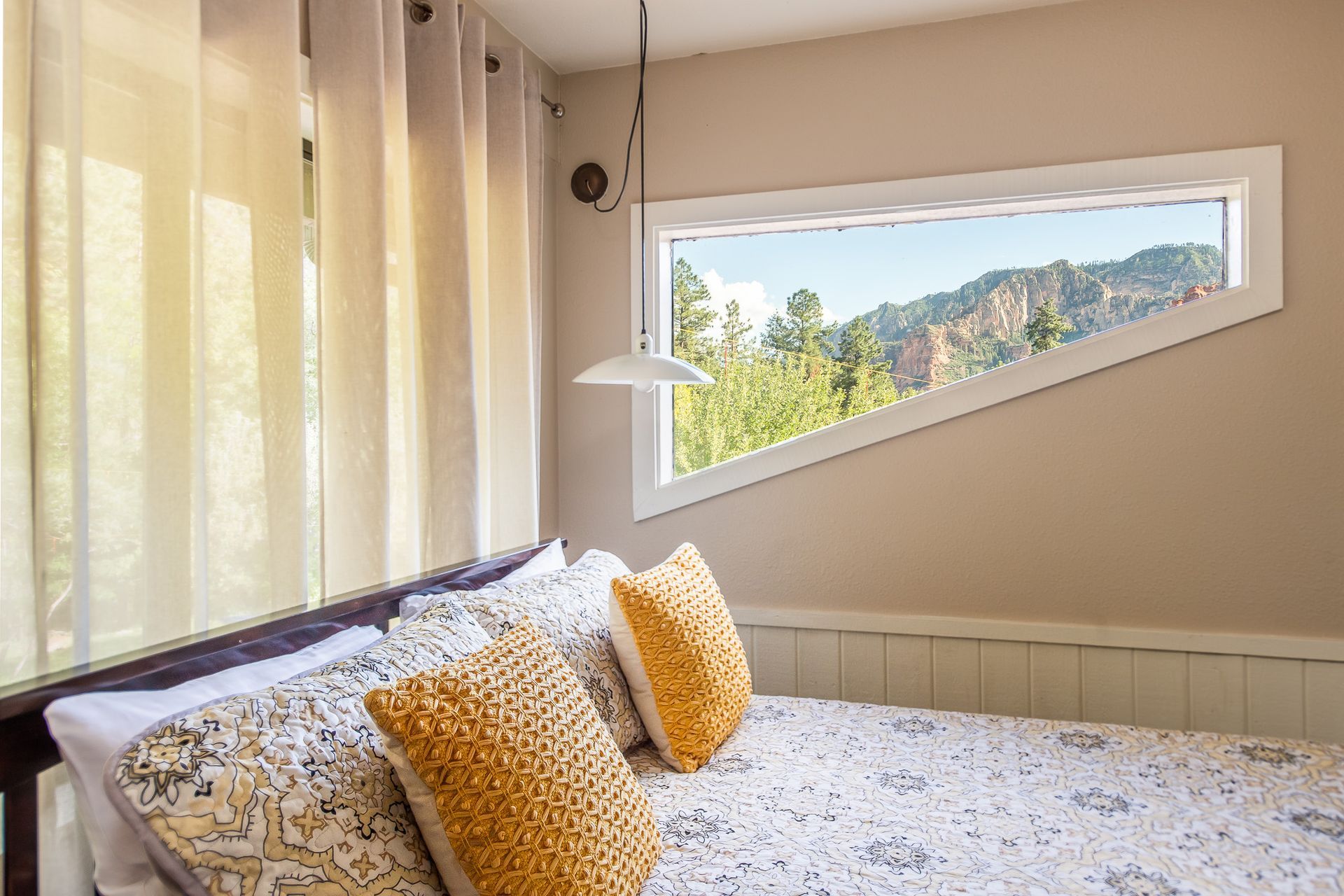 Bedroom with bed, geometric window framing a mountain view, cream-colored walls, and yellow throw pillows.