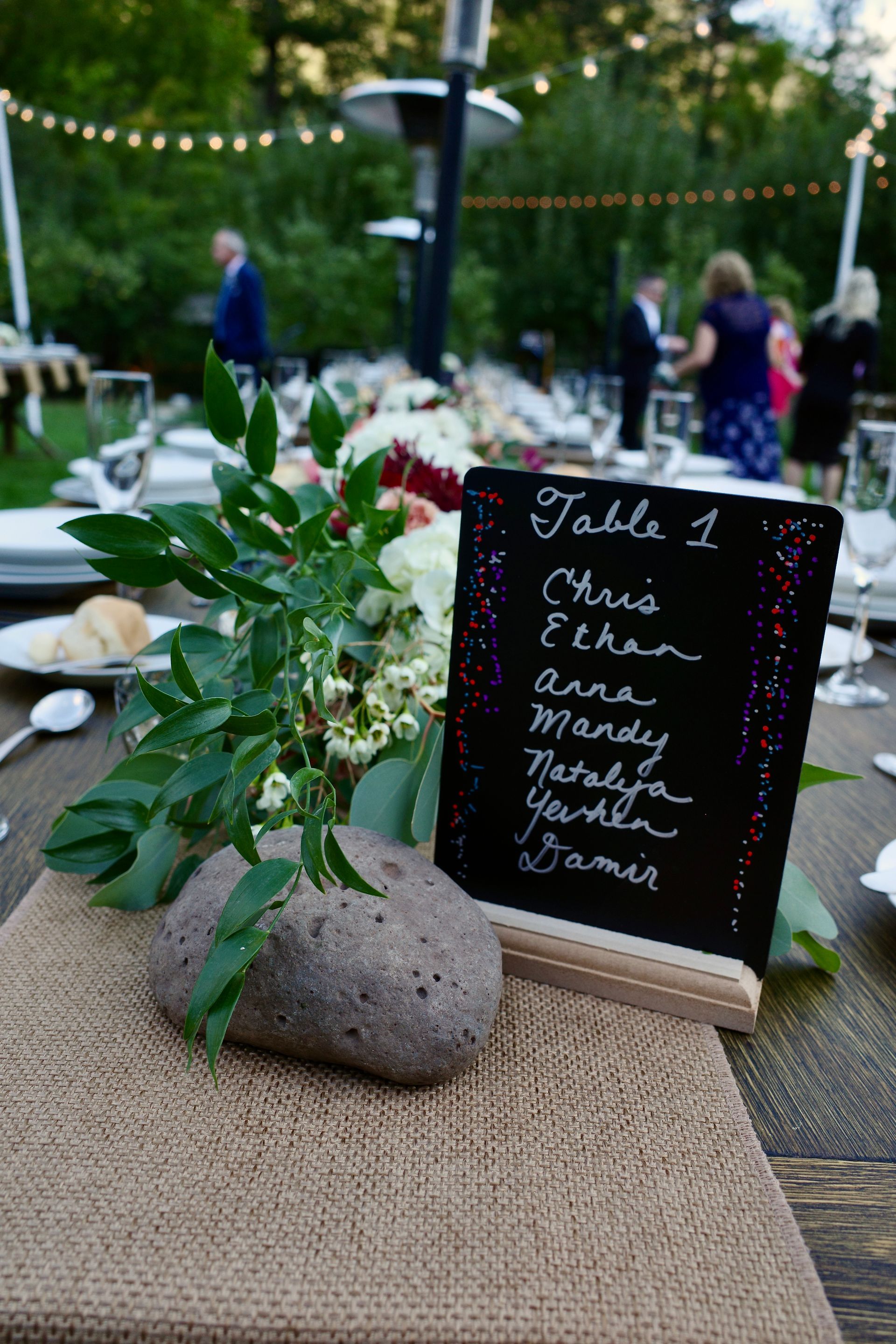 Table setting with guest names on a chalkboard, flower arrangement, and people in the background.