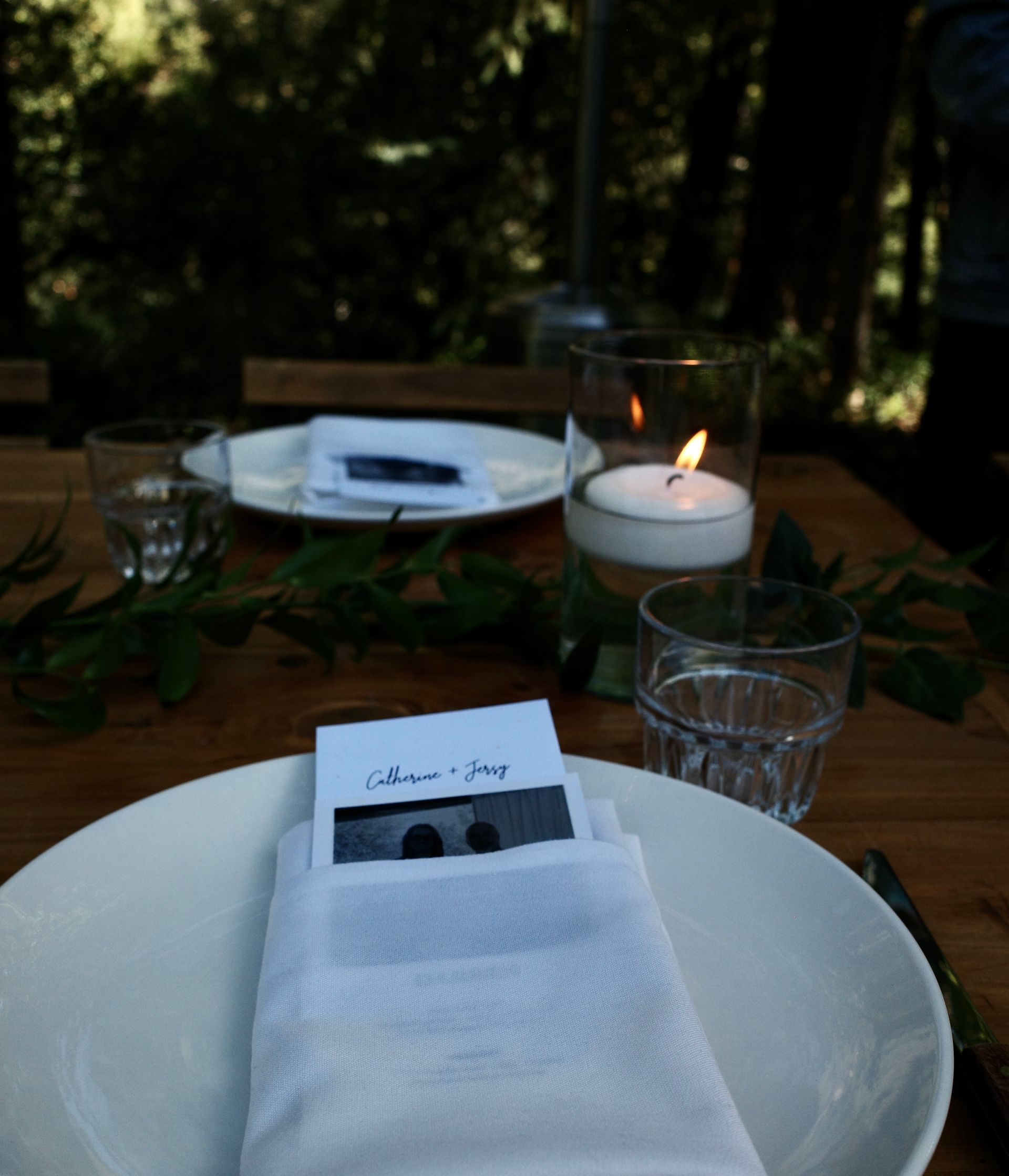 Table setting outdoors with a white plate, napkin, glass, candle, and a name card.