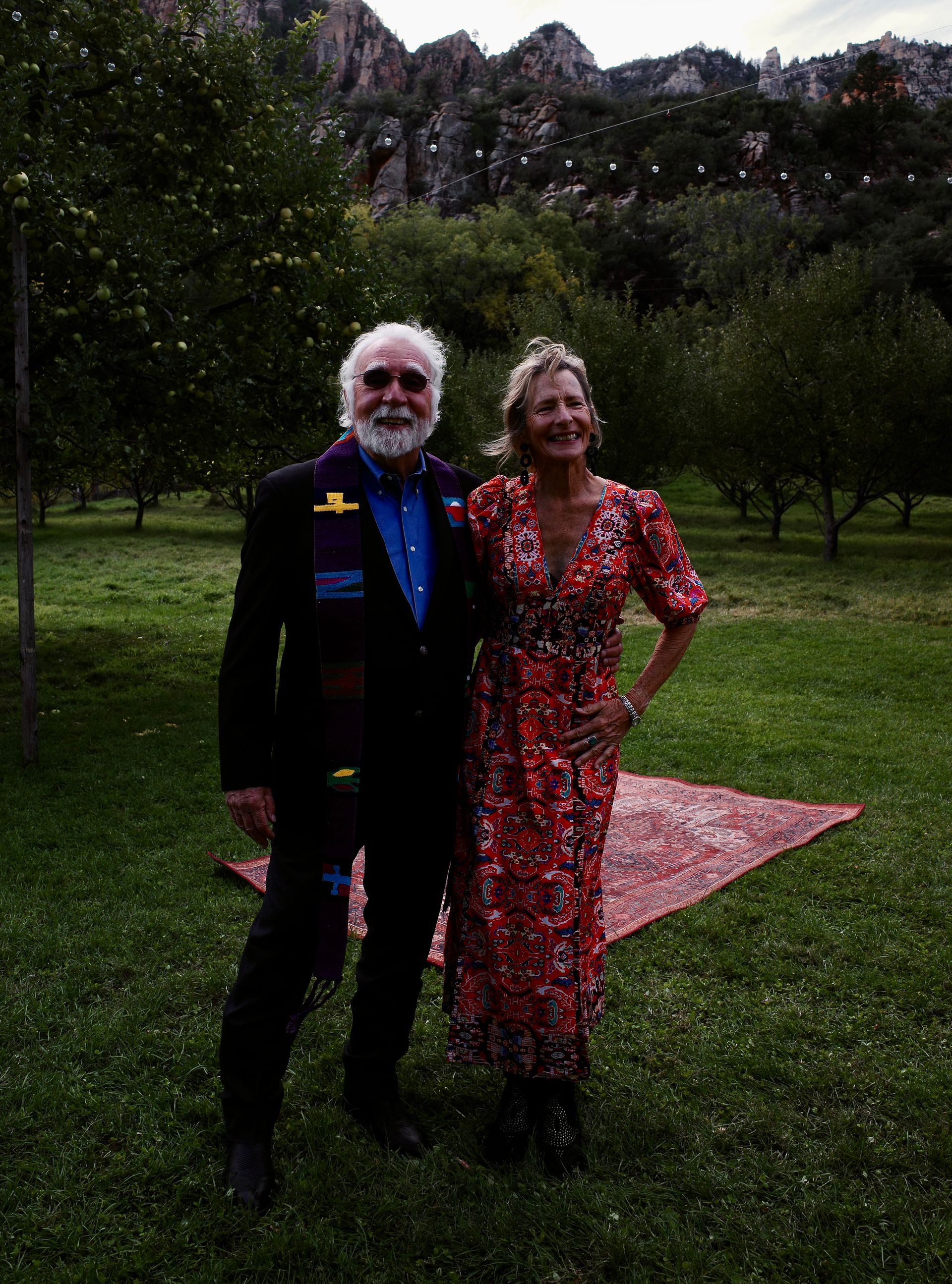 Man and woman posing outdoors in an orchard; man in suit, woman in floral dress, mountains in background.