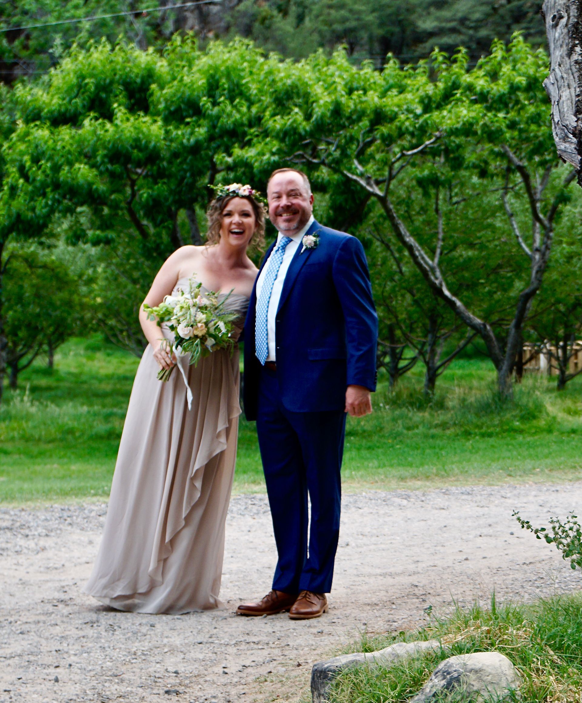 Wedding couple smiling outdoors; bride in beige dress, holding flowers; groom in blue suit.