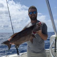 Man holding a large, dark fish on a boat, smiling, sunglasses, ocean background.