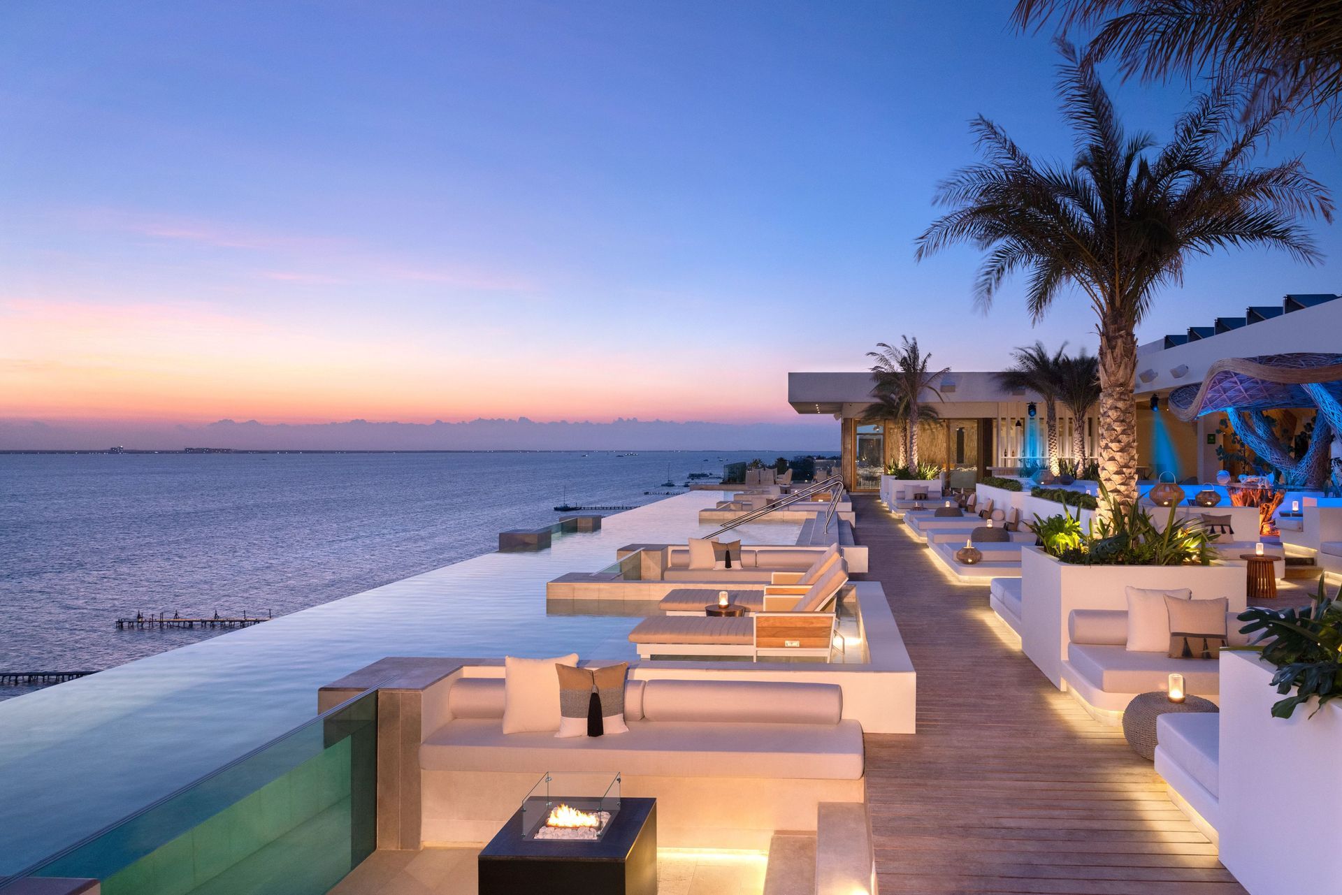 Luxury outdoor lounge with infinity pool overlooking the ocean at sunset. White furniture, palm trees.