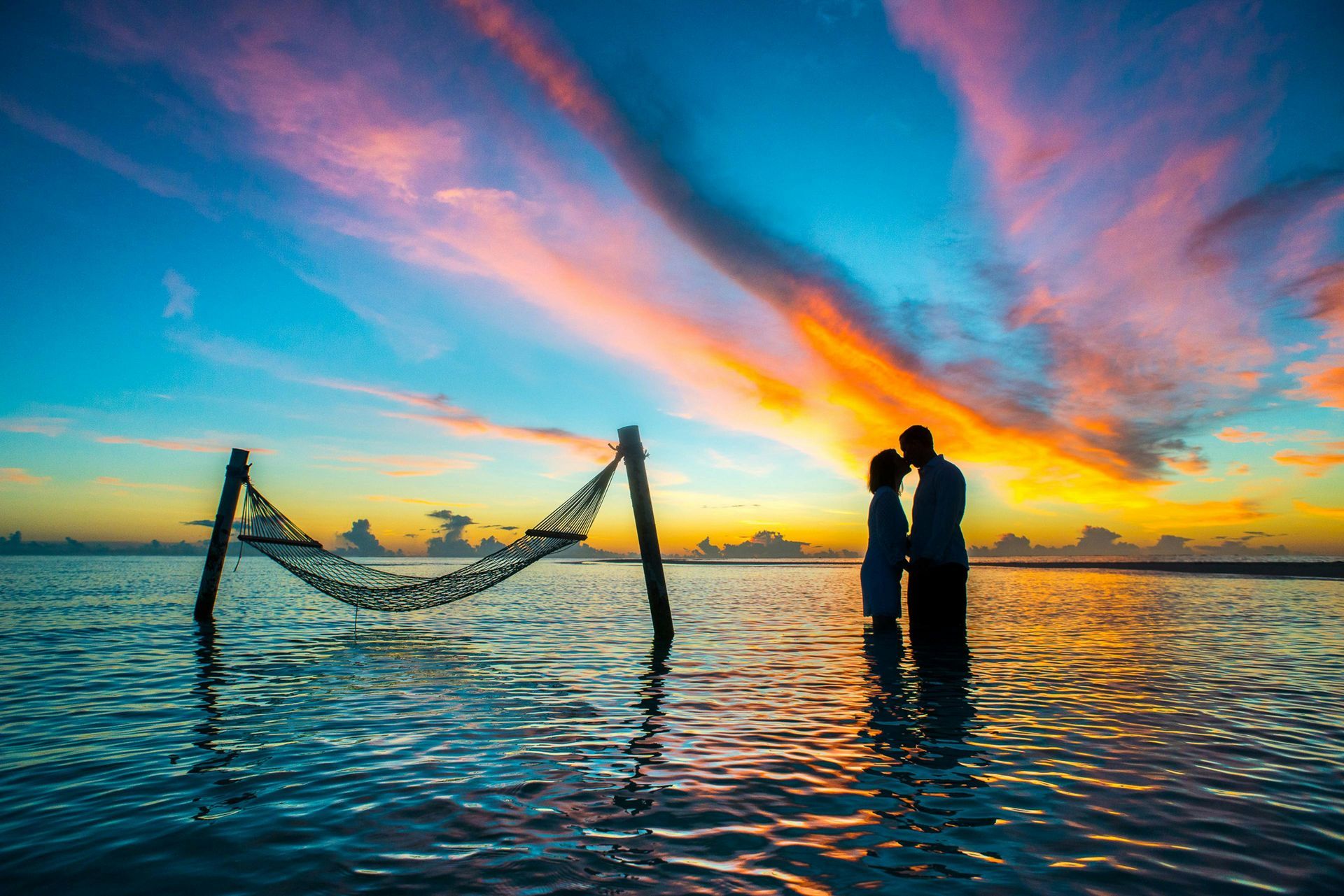 Couple silhouetted in shallow water, kissing at sunset, with a hammock nearby.