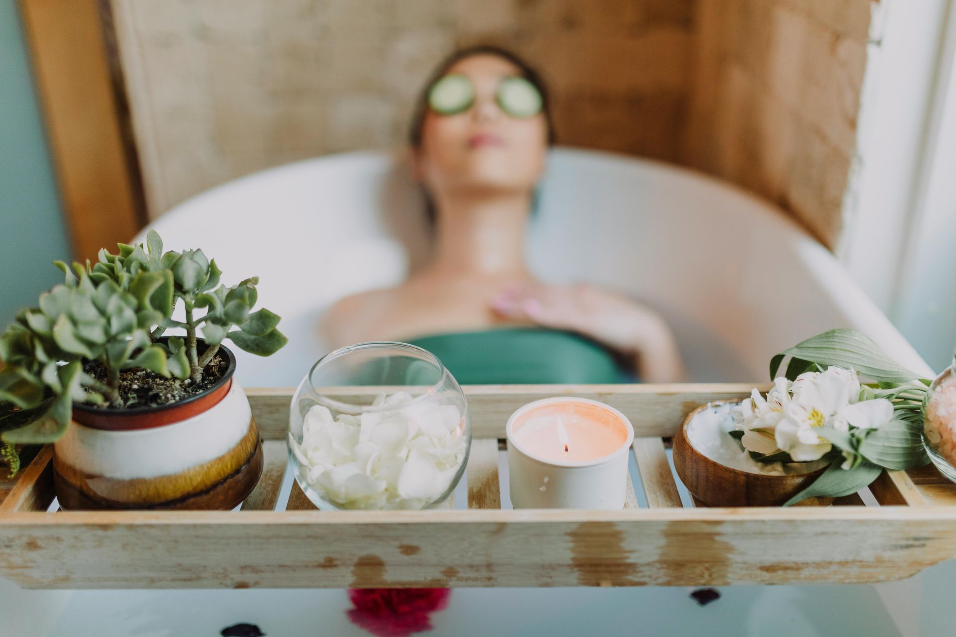 A woman is taking a relaxing bath with cucumber slices on her eyes and candle and plants on a tray above the bathwater.