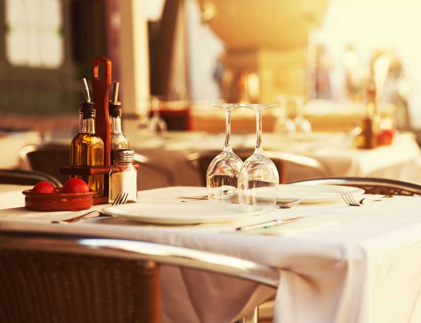 Tavolo da ristorante illuminato dal sole, apparecchiato con tovaglia bianca, bicchieri da vino, posate, bottiglie e mele rosse.