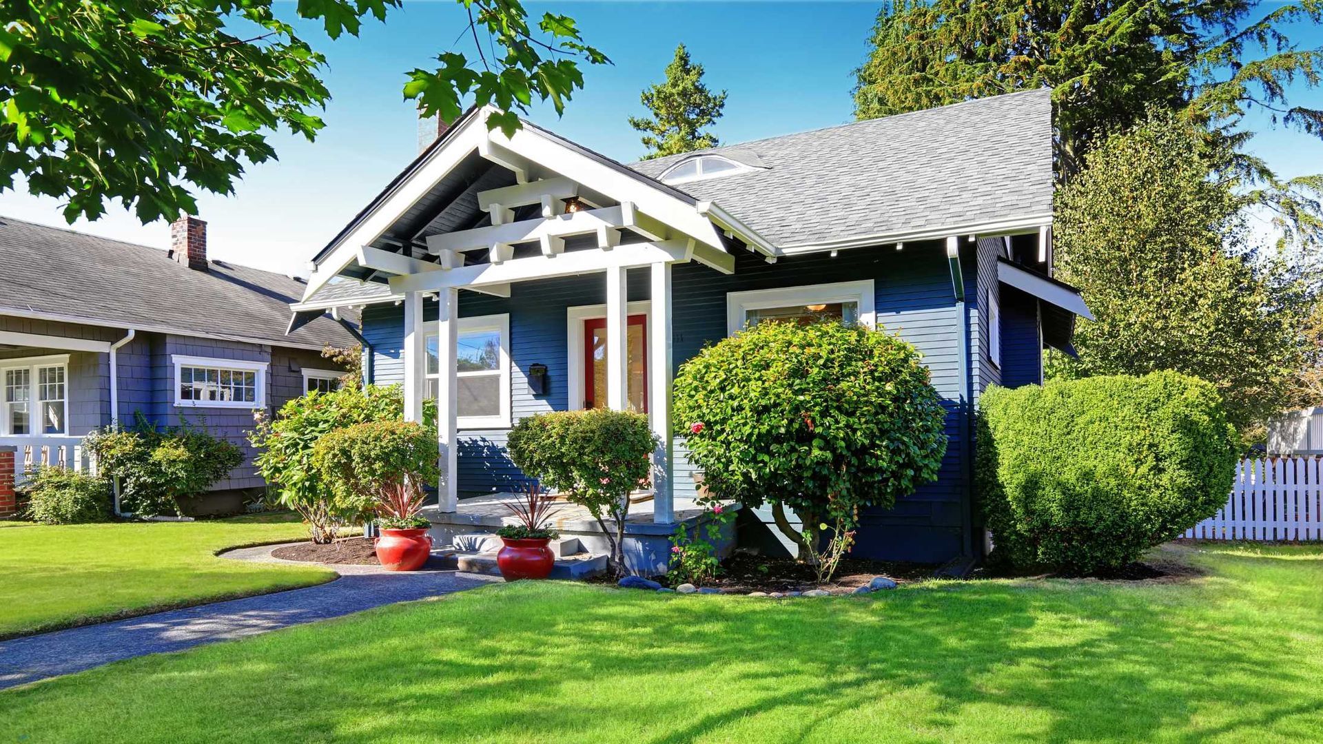 Blue bungalow house with white trim, front porch, and manicured green lawn on a sunny day