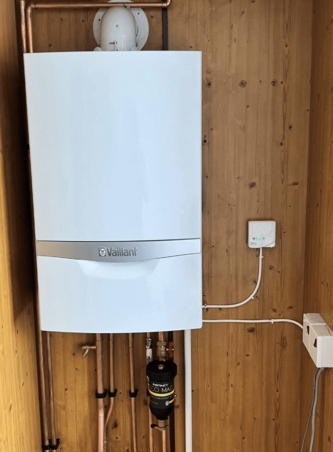 A white boiler is hanging on a wooden wall next to copper pipes.