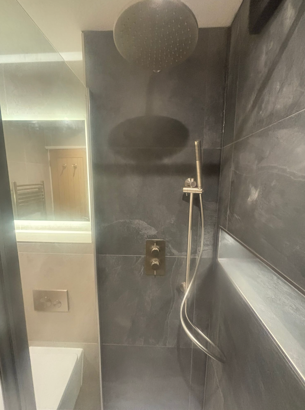 Dark gray tiled shower with round showerhead, handheld shower, and control panel. Mirror to the left.