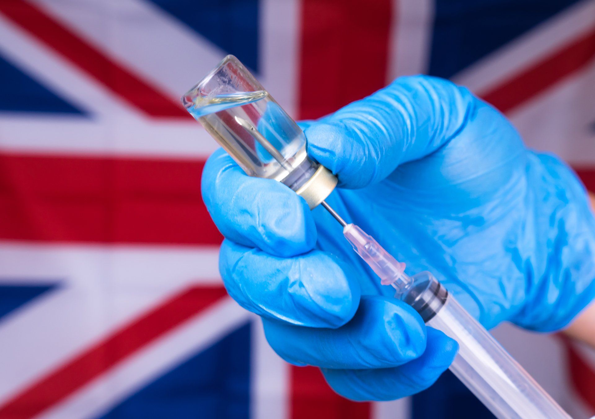 A person wearing blue gloves is holding a syringe and a bottle of vaccine in front of a british flag.