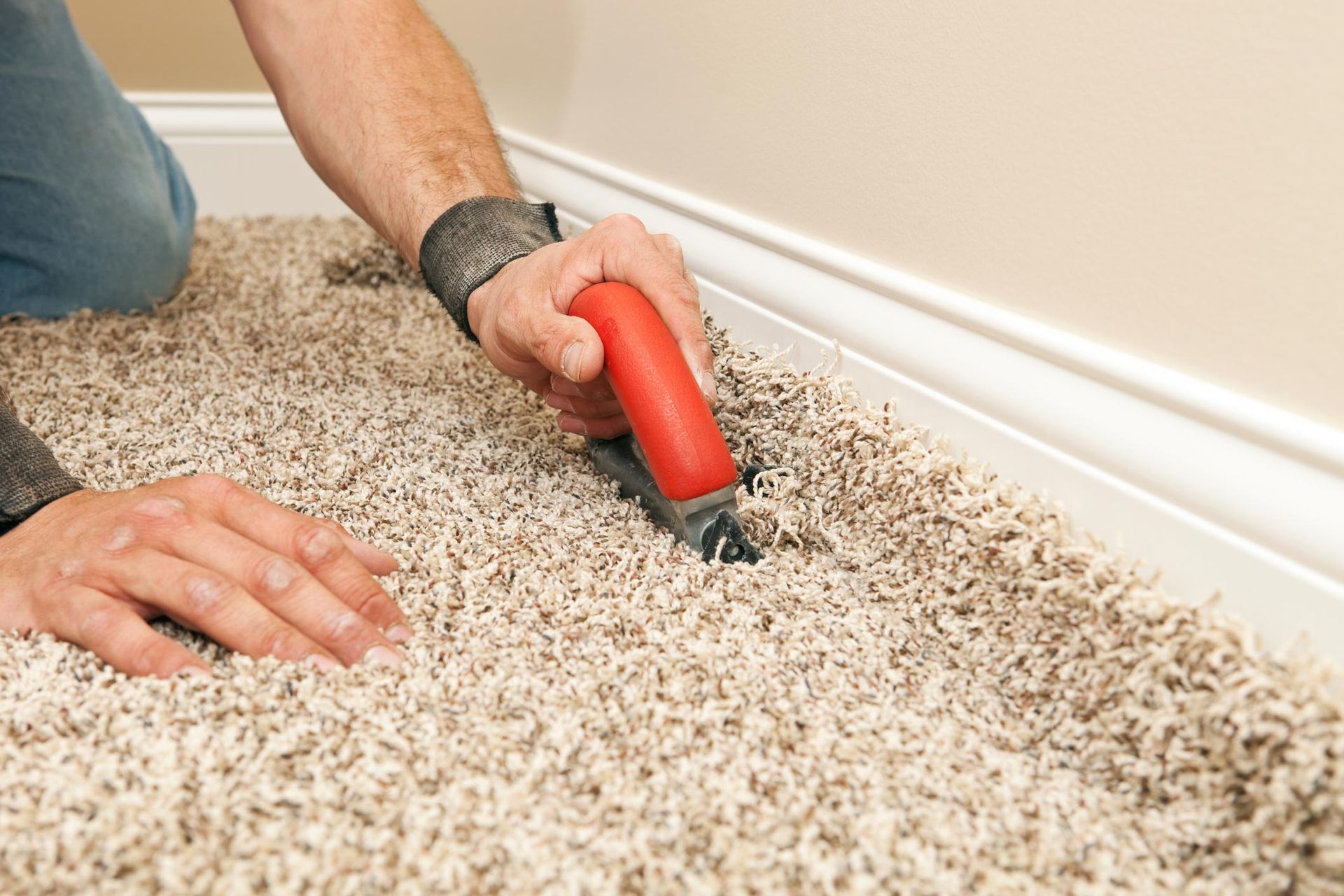 Person cuts carpet edge with red-handled tool near white baseboard.