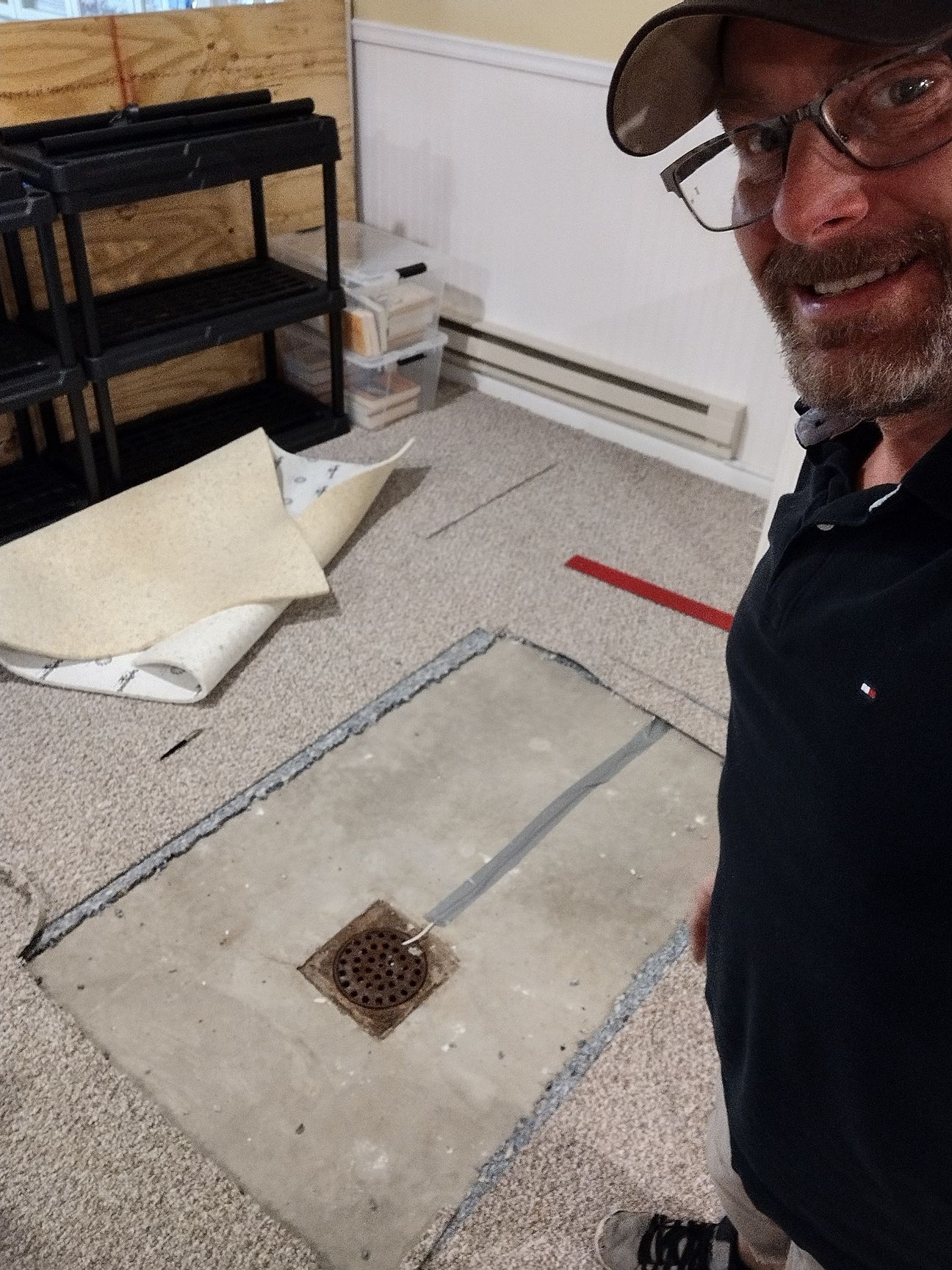 Man smiles while standing in a room with concrete floor cutout around a drain. Beige carpet, storage shelves visible.