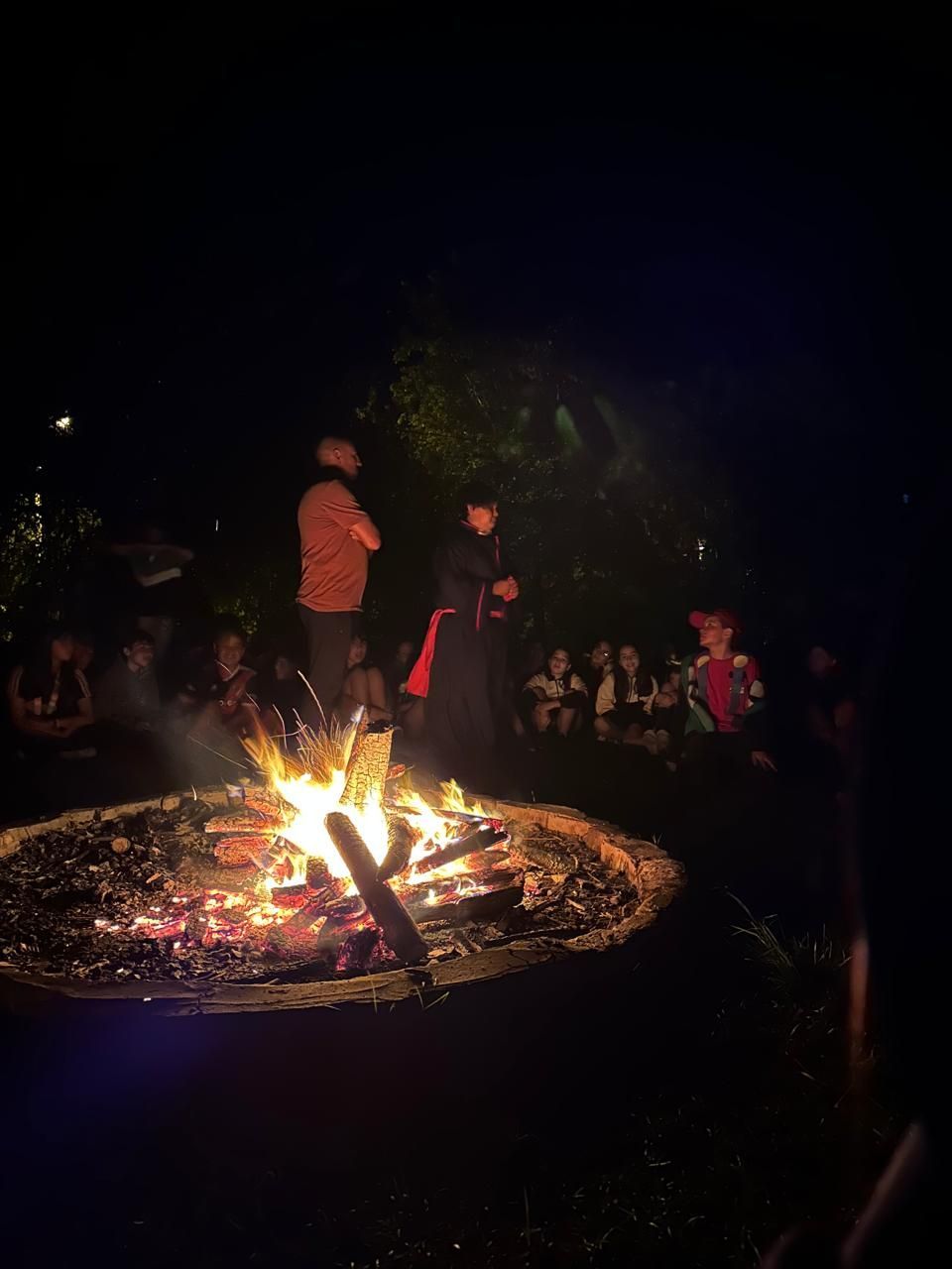 Un grupo de personas está de pie alrededor de una fogata por la noche.