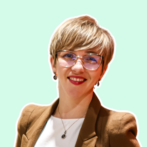 A woman wearing glasses and a brown jacket is smiling.