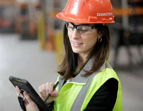 Two construction workers are standing next to a car in a warehouse looking at a tablet.