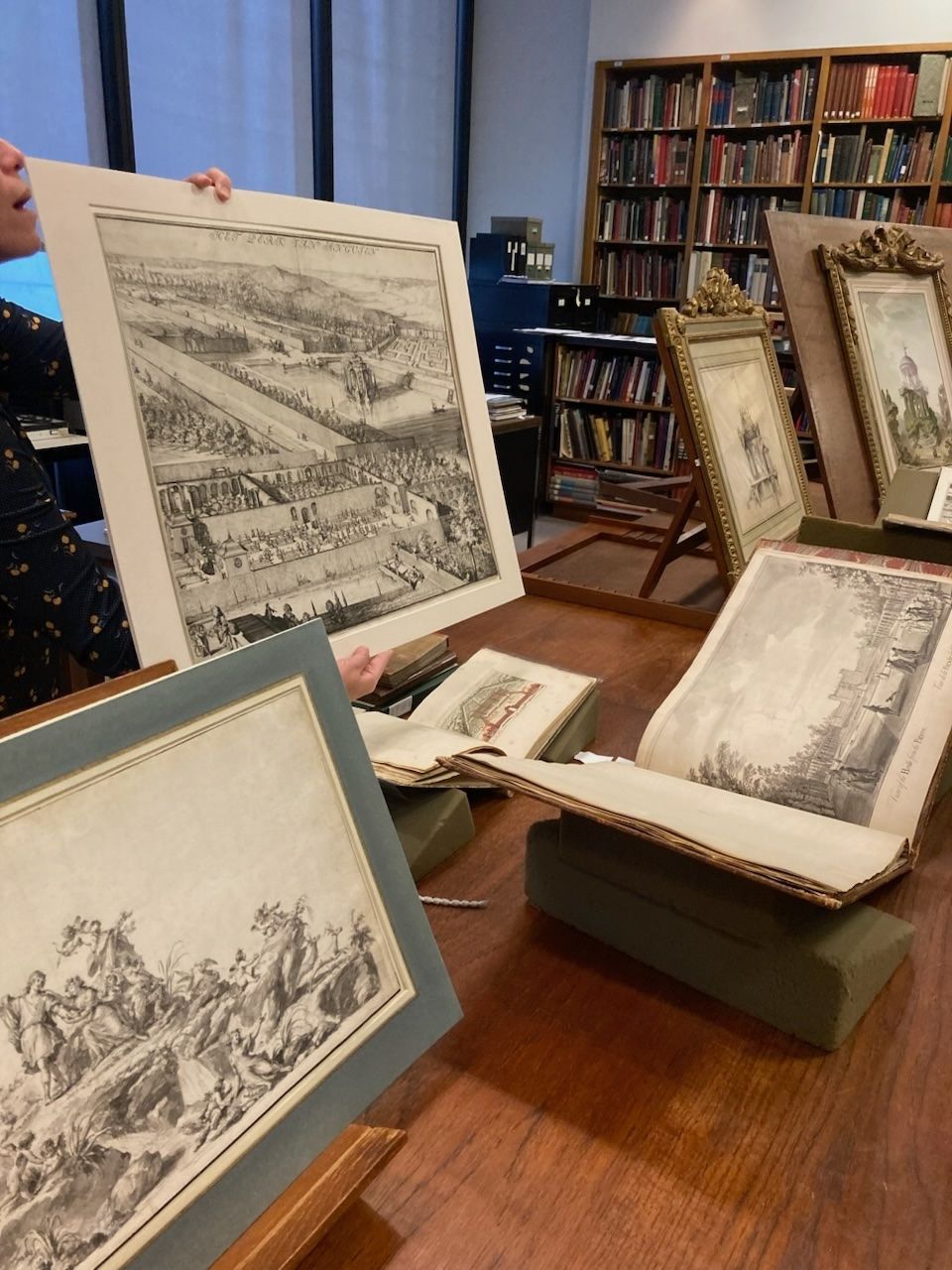 Person holding a large print in a library, surrounded by displayed artworks and books on shelves.
