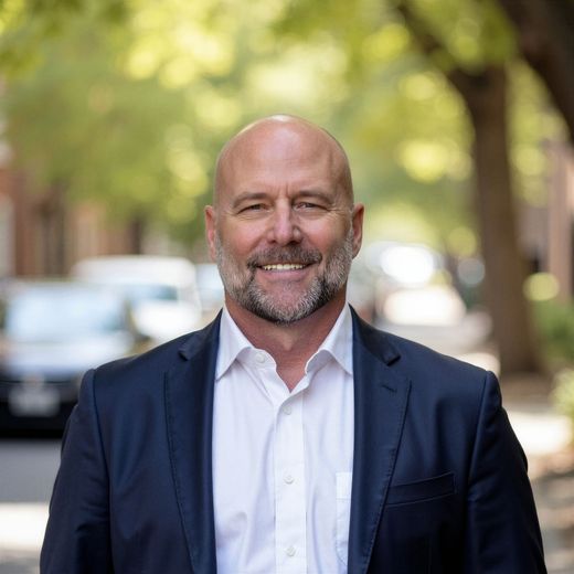 Smiling bald man in a navy blazer and white shirt standing on a tree-lined street.