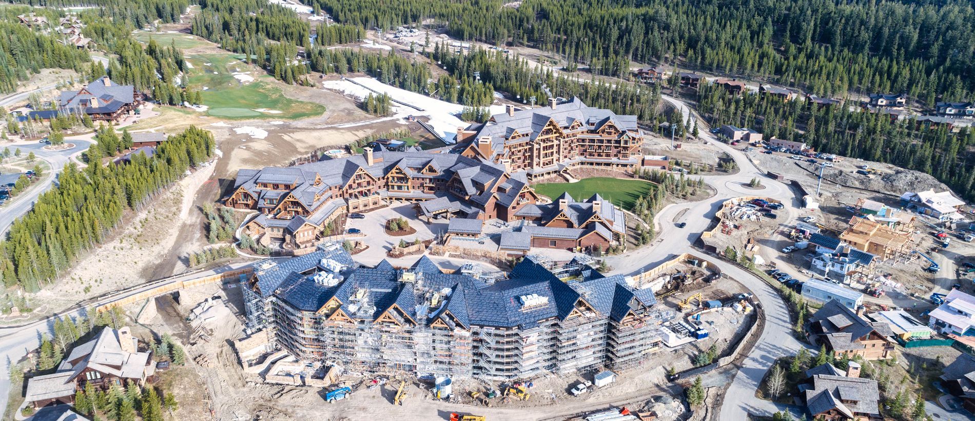 Aerial view of a large mountain resort under construction, with multiple buildings and forested hills nearby
