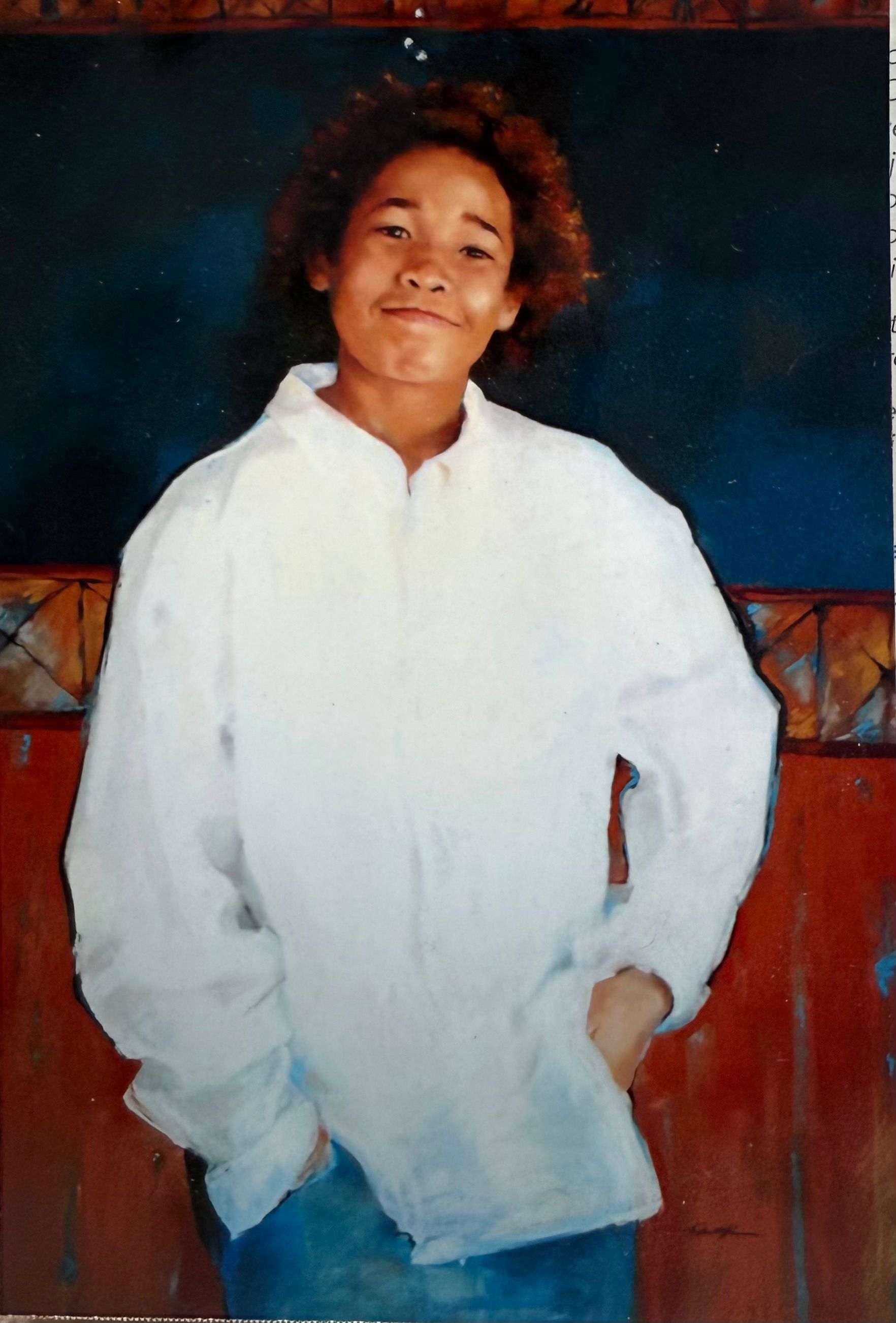 Young person in white shirt, hands in pockets, smiling. Standing in front of red and blue wall.