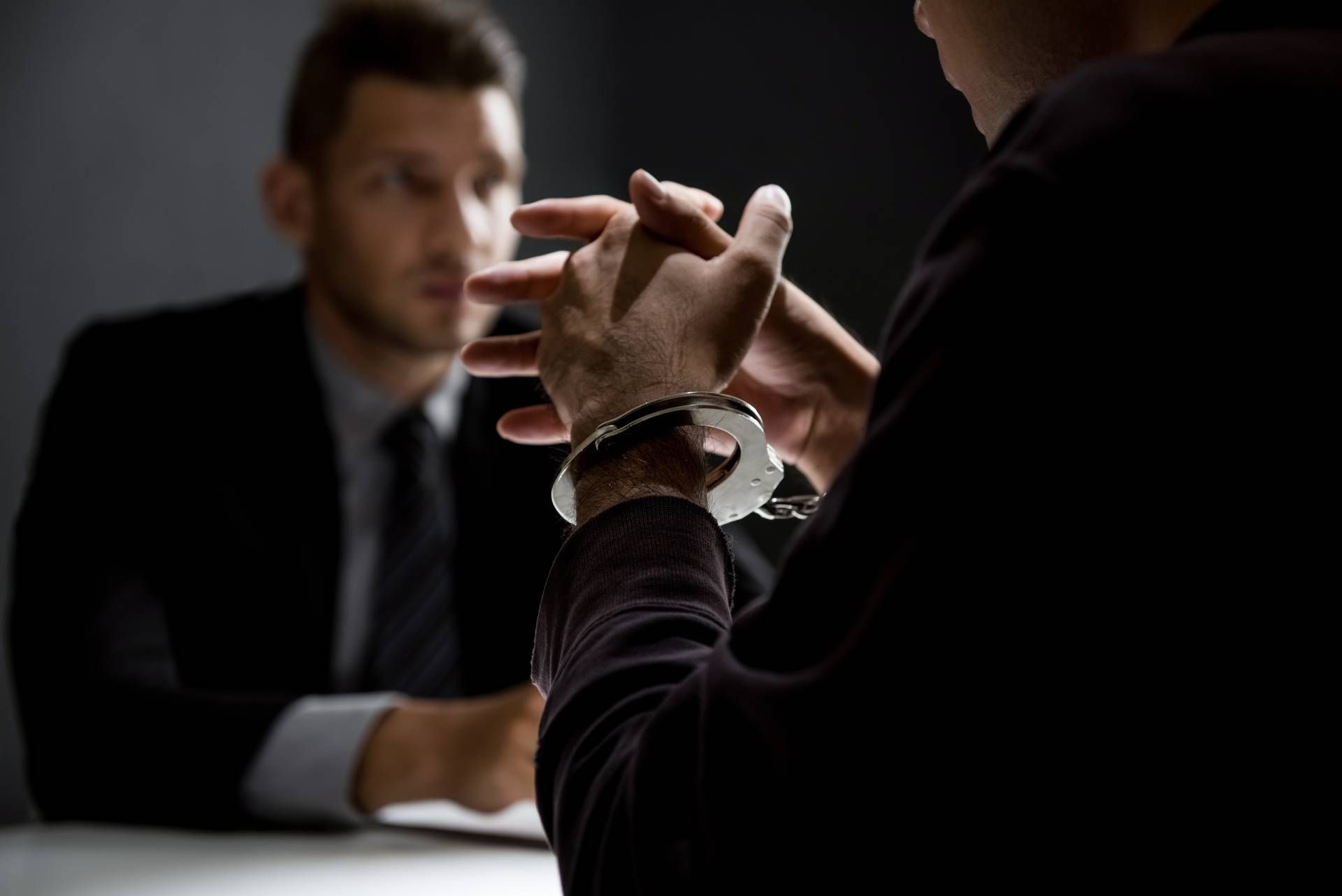 Handcuffed person facing an investigator, in a dimly lit interrogation room.